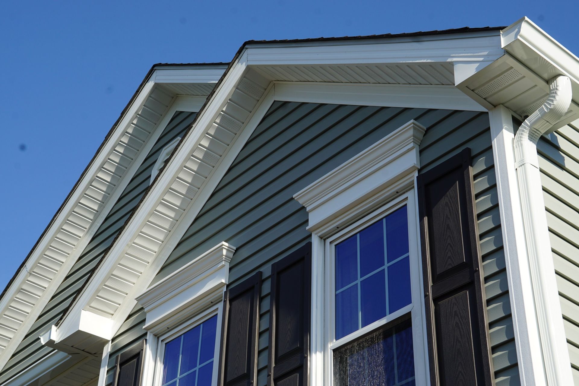 Close-up of a house exterior with light green siding, white trim, black shutters, and blue windows against a clear blue sky.