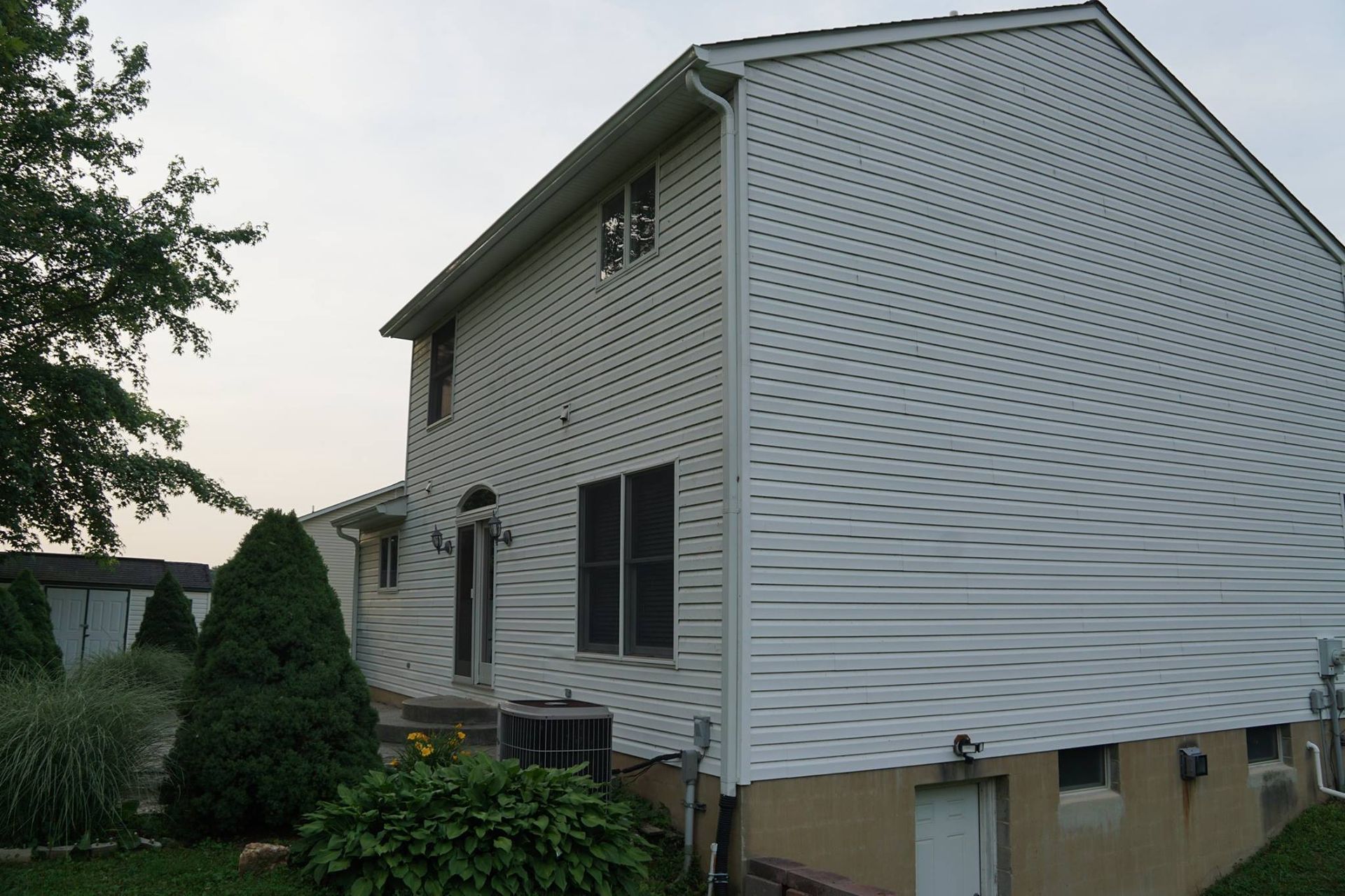 Side view of a two-story white-sided house with a lawn and shrubbery.