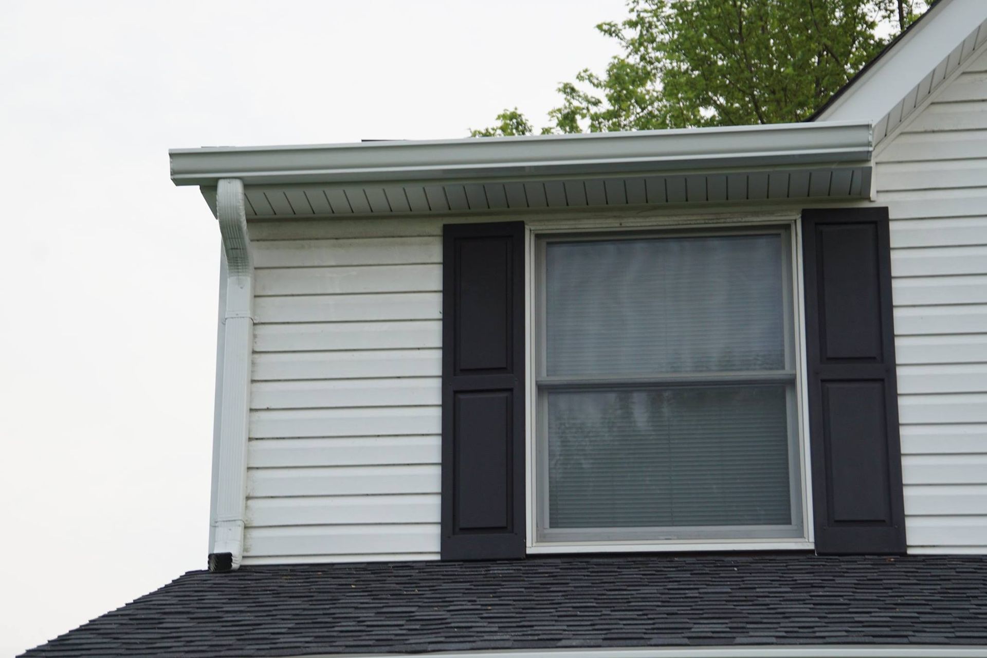 White house siding with a window, black shutters, and a dark roof. Gutters run along the roofline.