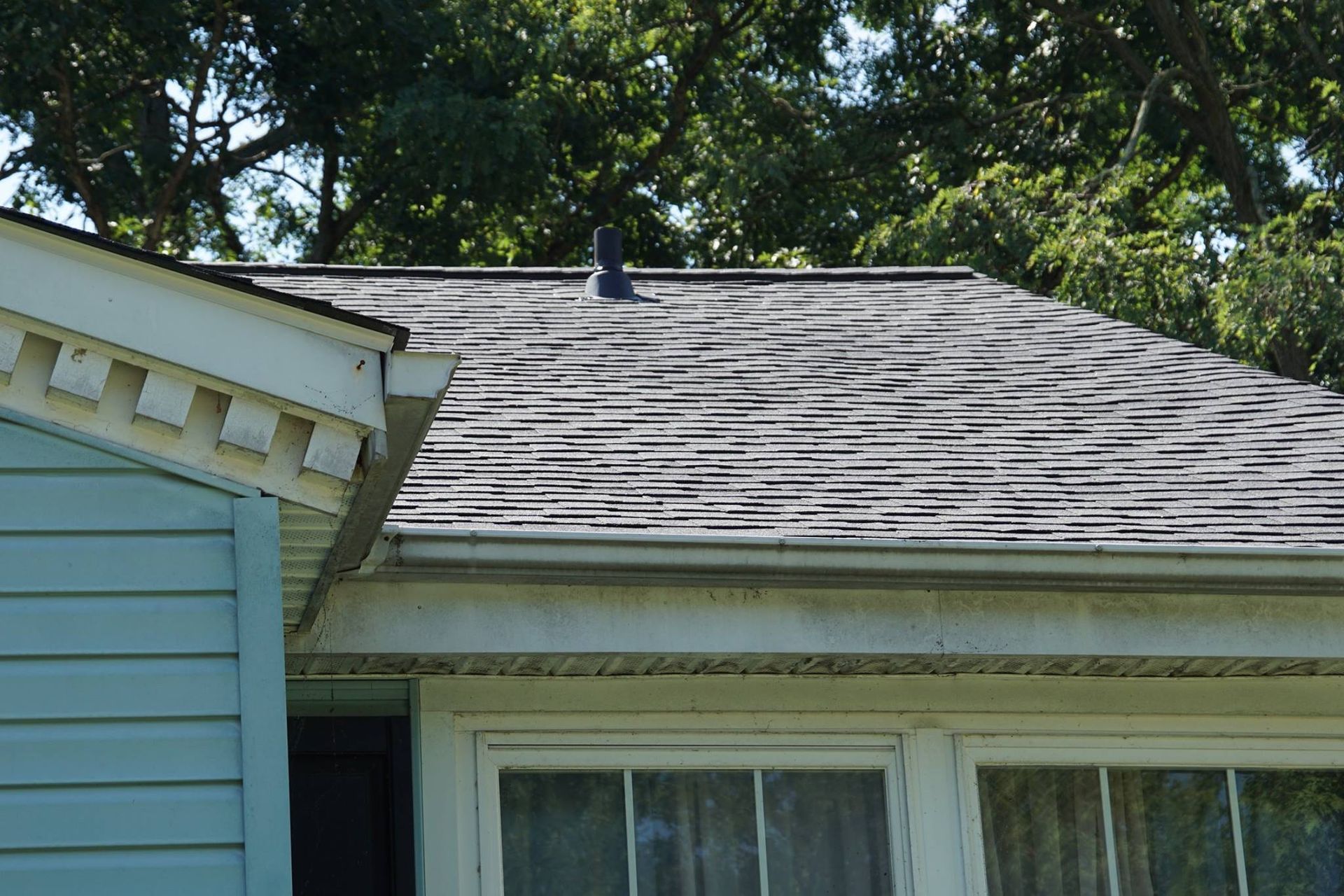 Roof of a light blue house with weathered grey shingles and a small, dark vent. Green trees in background.