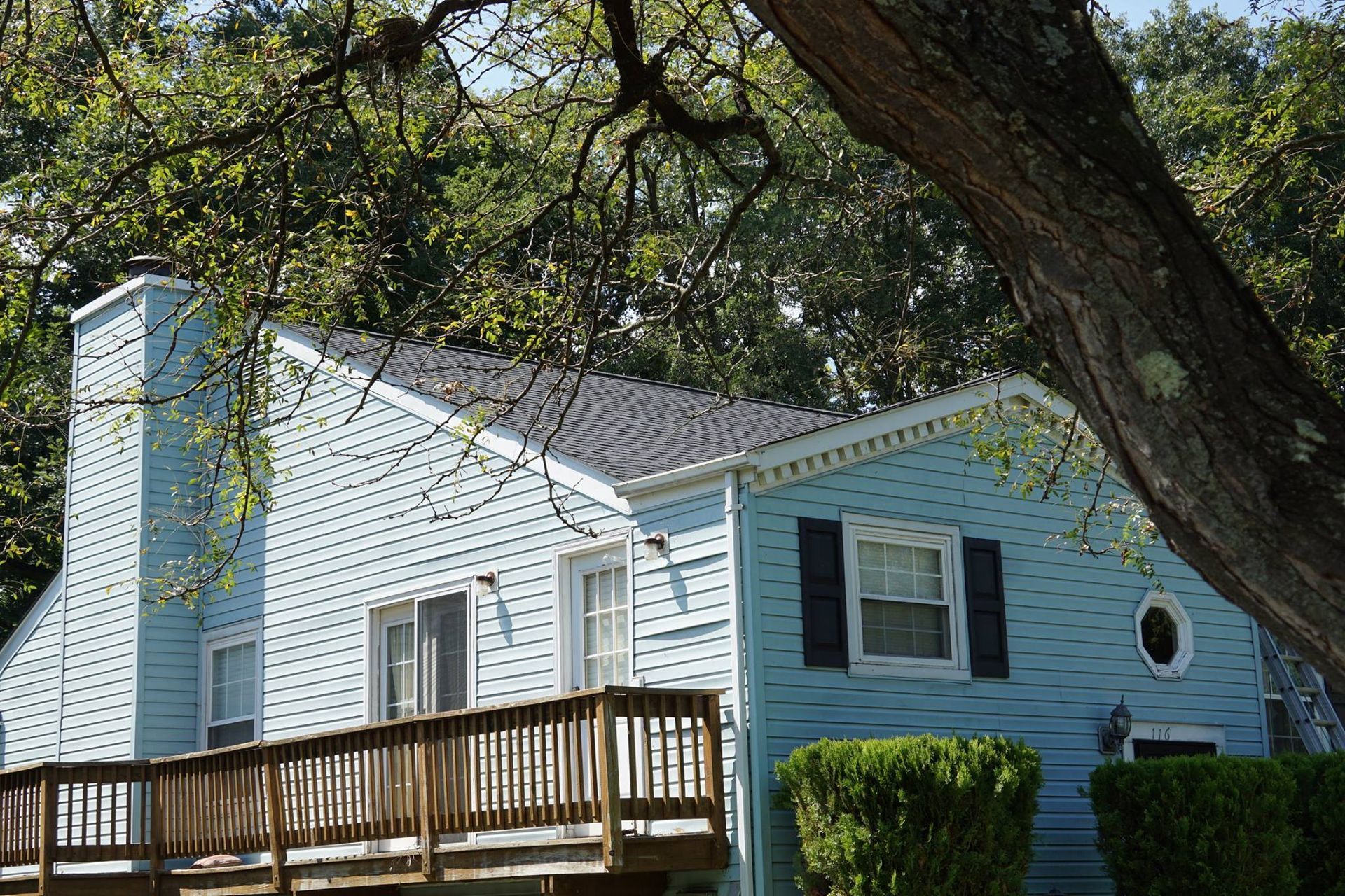 Light blue house with black shutters, chimney, and wooden deck, framed by a tree.