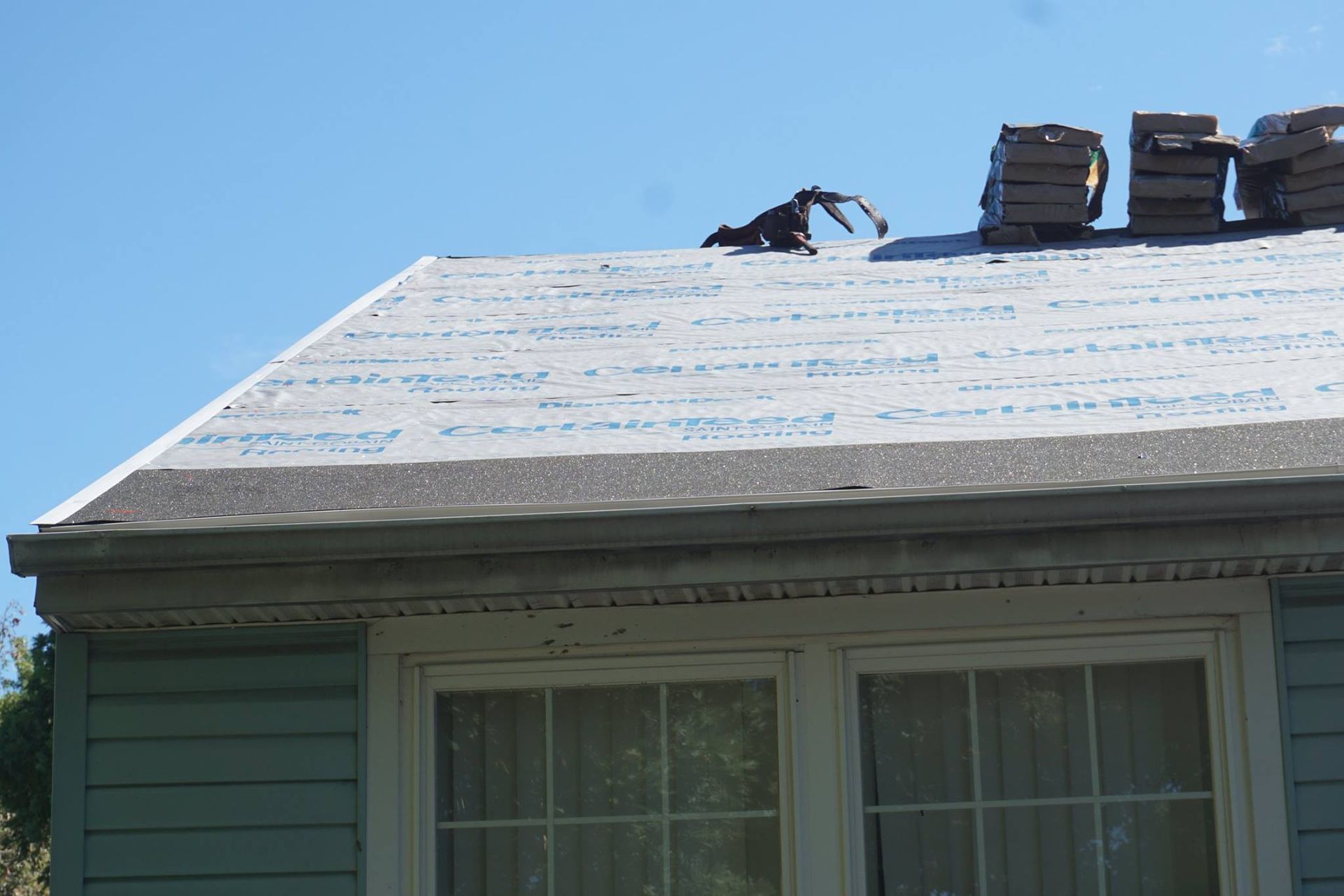 Person working on a roof with new underlayment; shingles stacked nearby, blue sky.