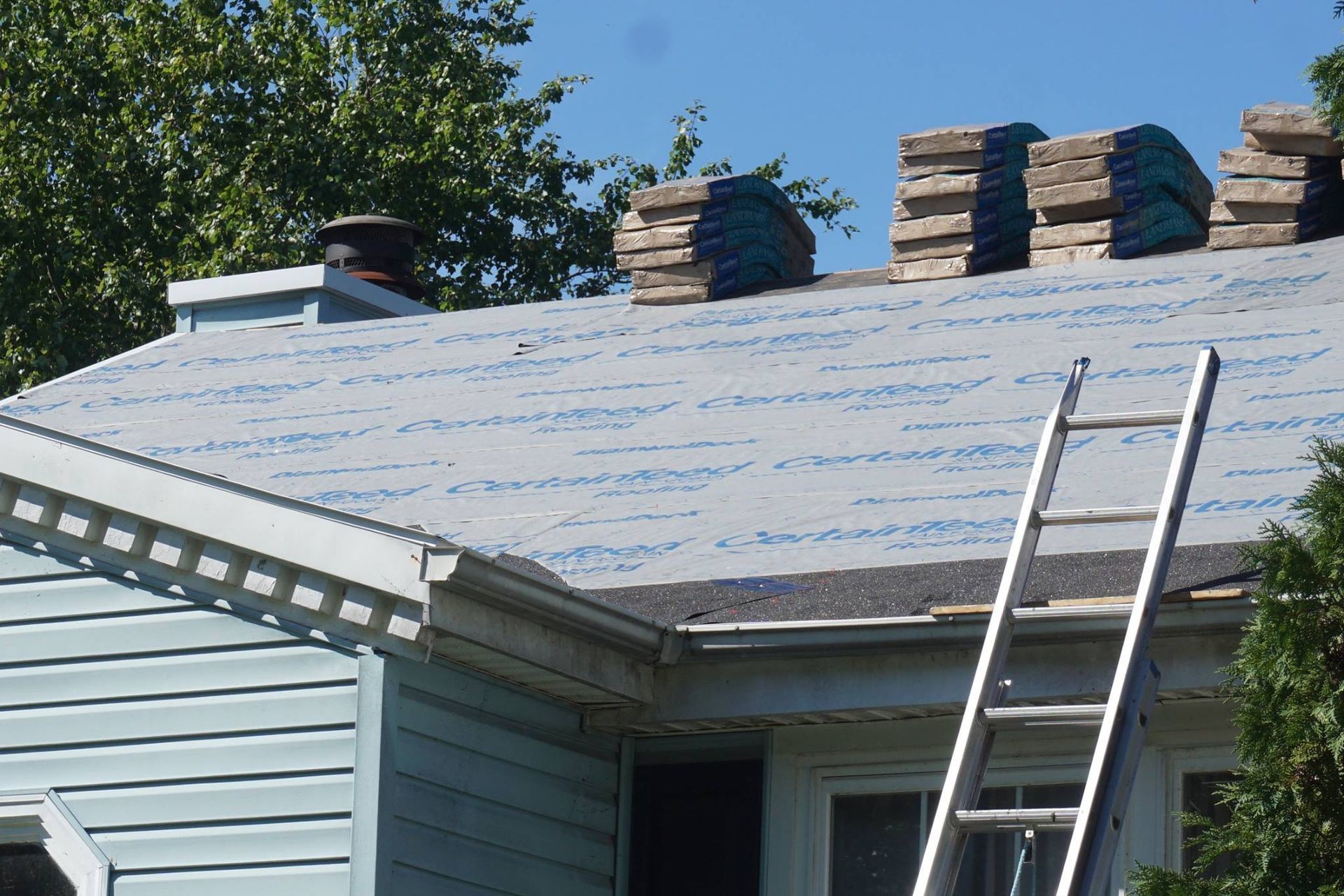 Roof replacement in progress: shingles stacked, ladder on roof, blue underlayment, and blue siding.