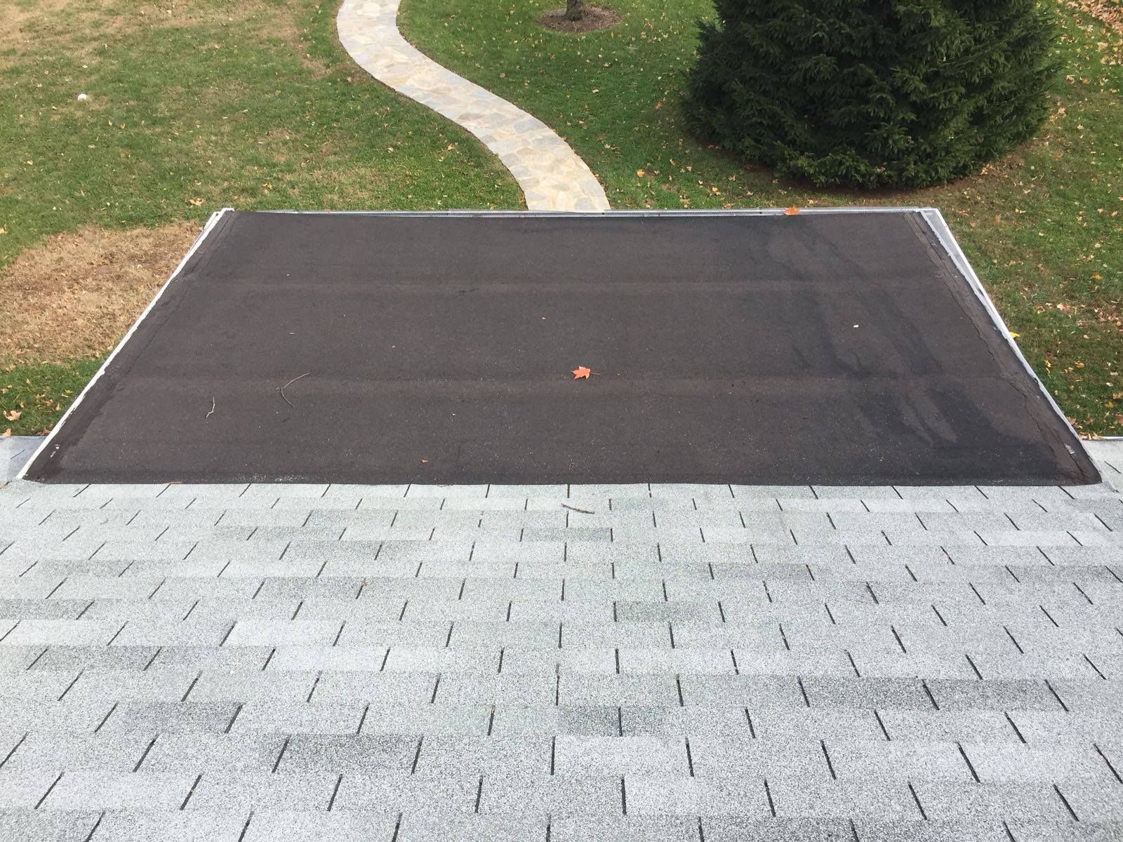 A section of dark roofing material over a light-colored shingle roof, surrounded by grass and a walkway.