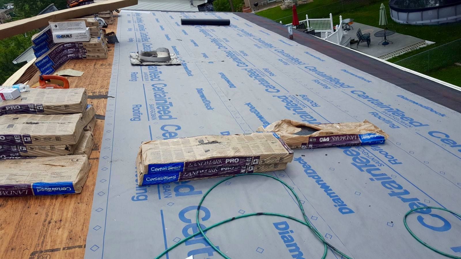 Roofing materials on a flat roof, including shingles, rolls of underlayment, and roof vents, outdoor setting.