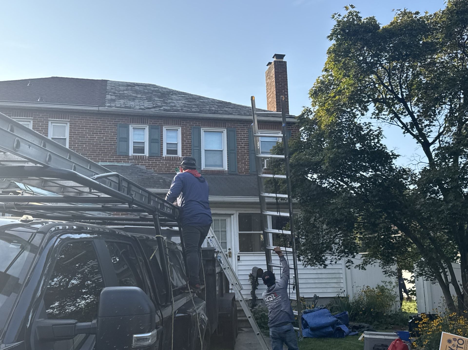 Two people working on a house roof. One on the truck rack, the other climbing a ladder. Bright sky.