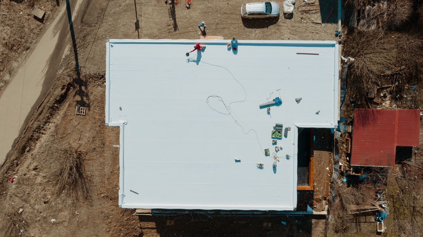 Overhead view of workers on a white roof, scattered tools, and a red-roofed building nearby.