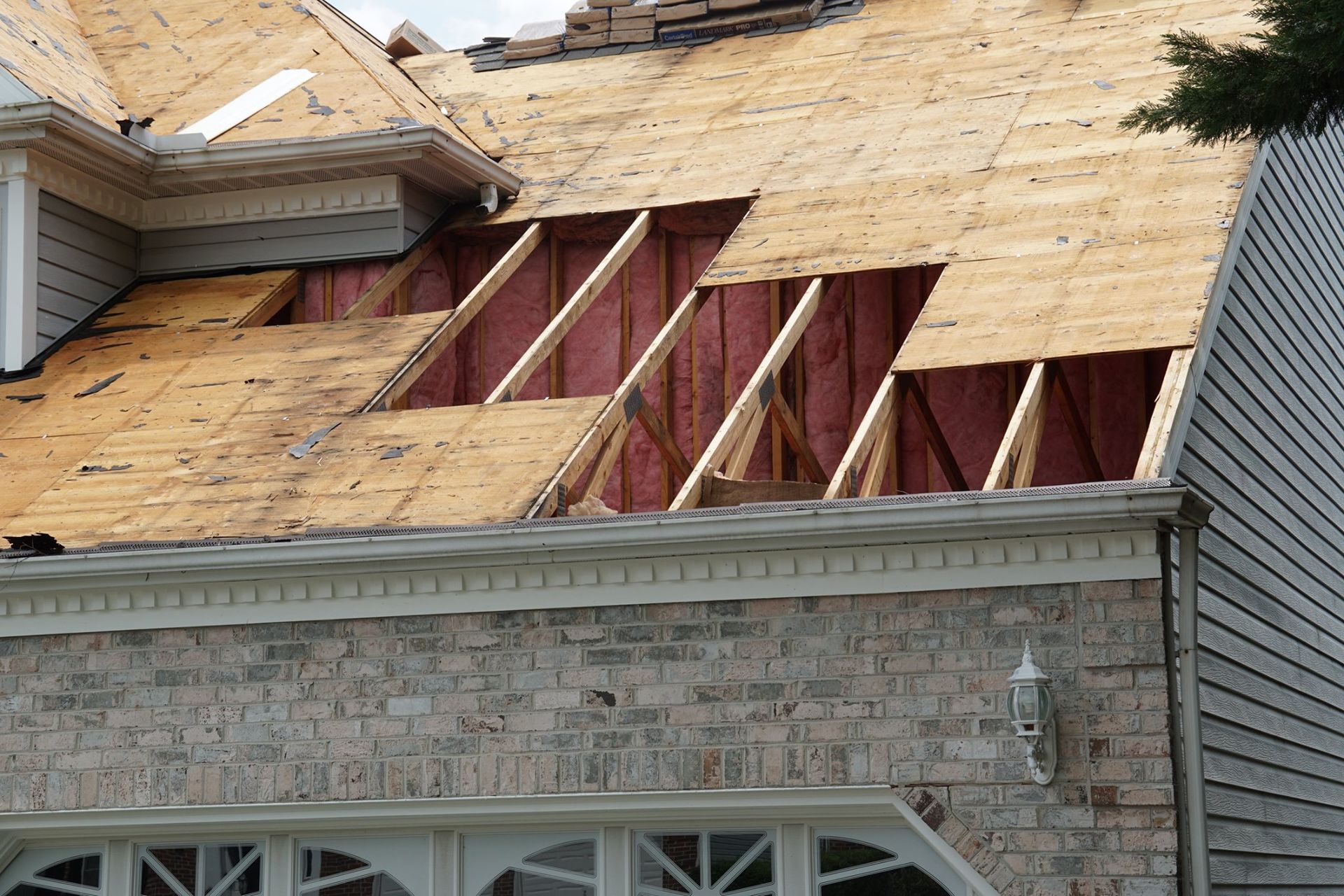 Roof with missing shingles revealing wood framing and pink insulation. Brick and siding walls visible.