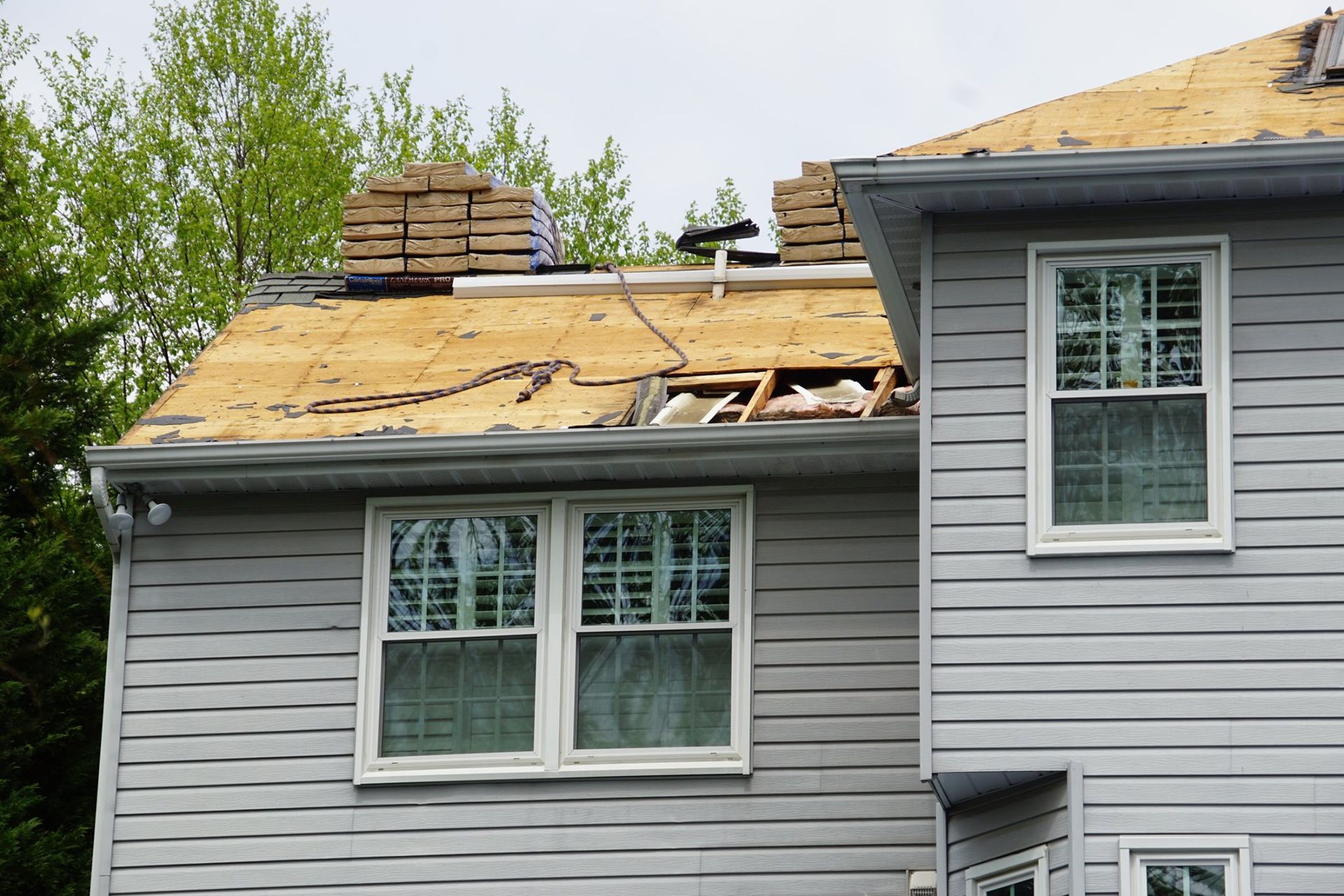 House roof partially torn off, showing exposed wood, with new shingles stacked on top. Gray siding.