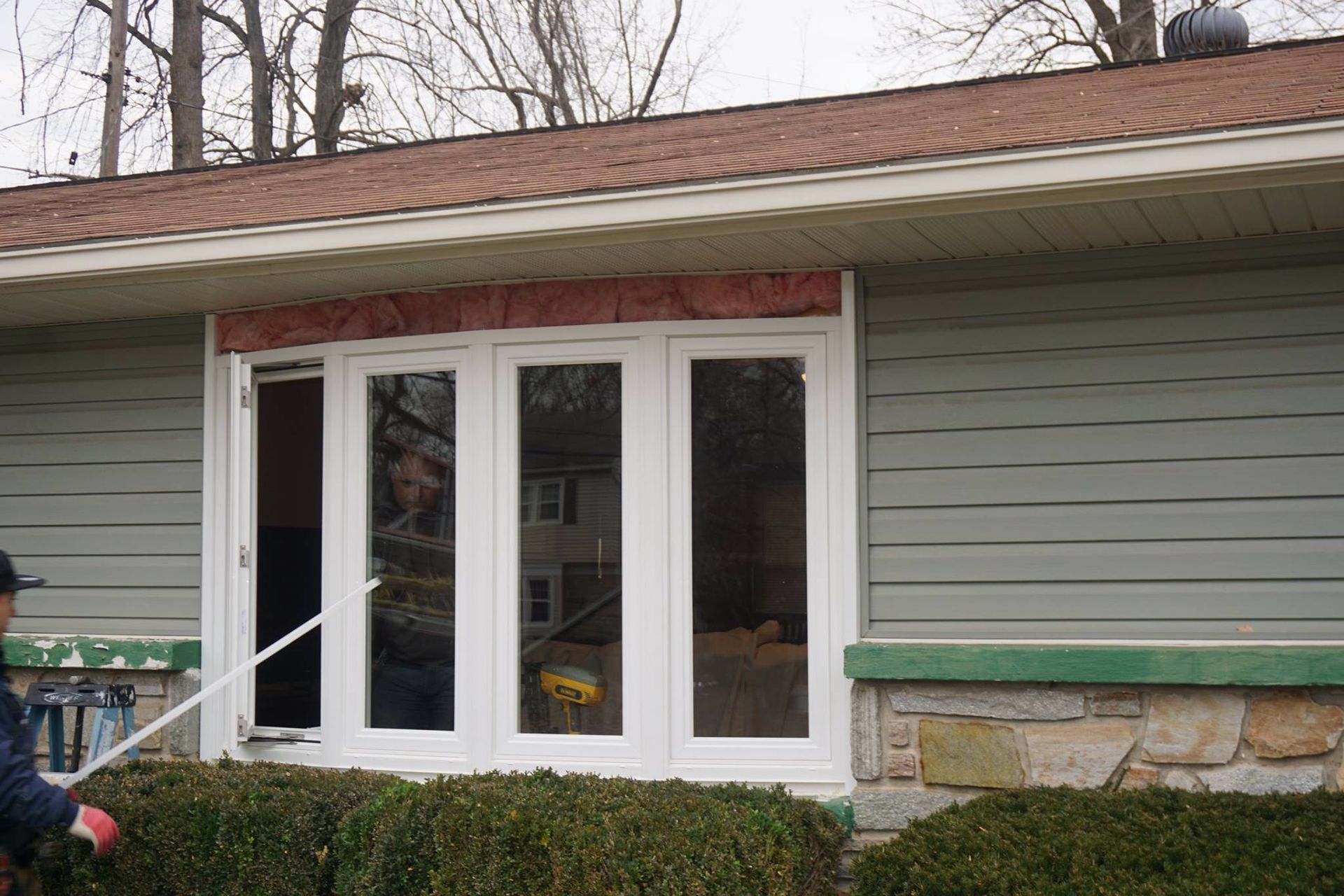Person cleaning window of a house with green siding, bushes in front.
