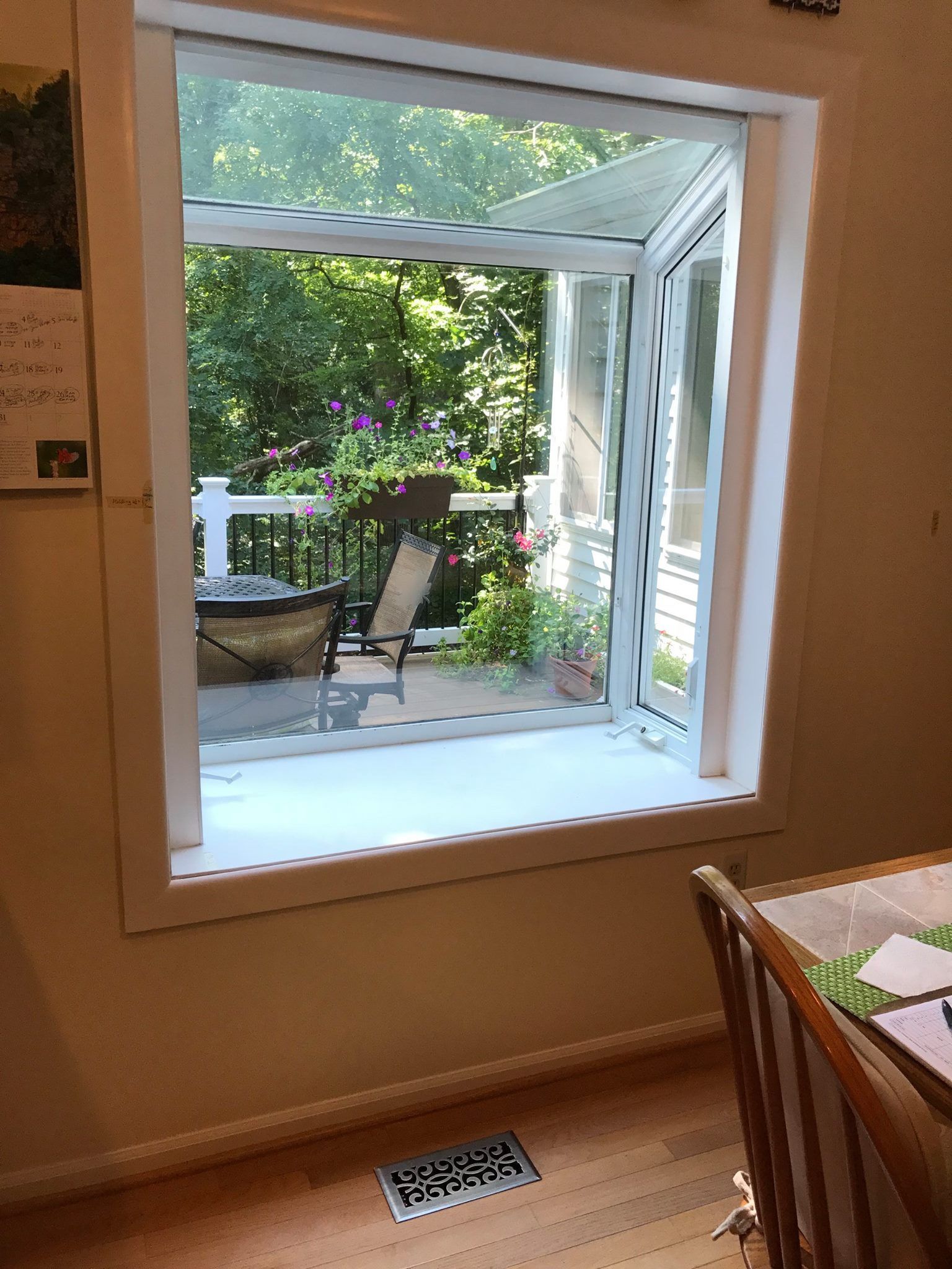Window with a view of a deck and greenery. White trim, wood floor, and a glimpse of a dining table.