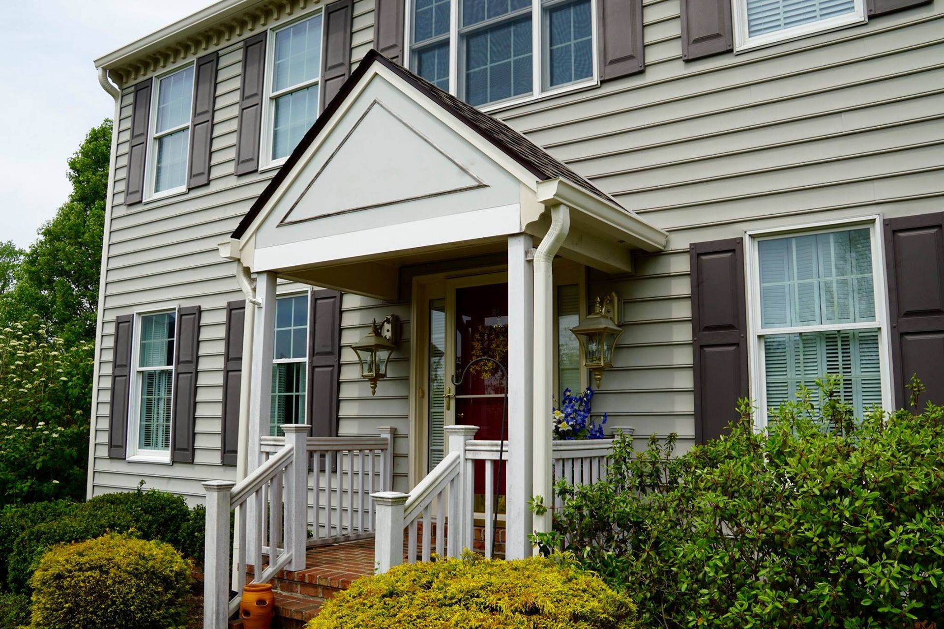 Beige house with a small porch and red door; brown shutters and green bushes.