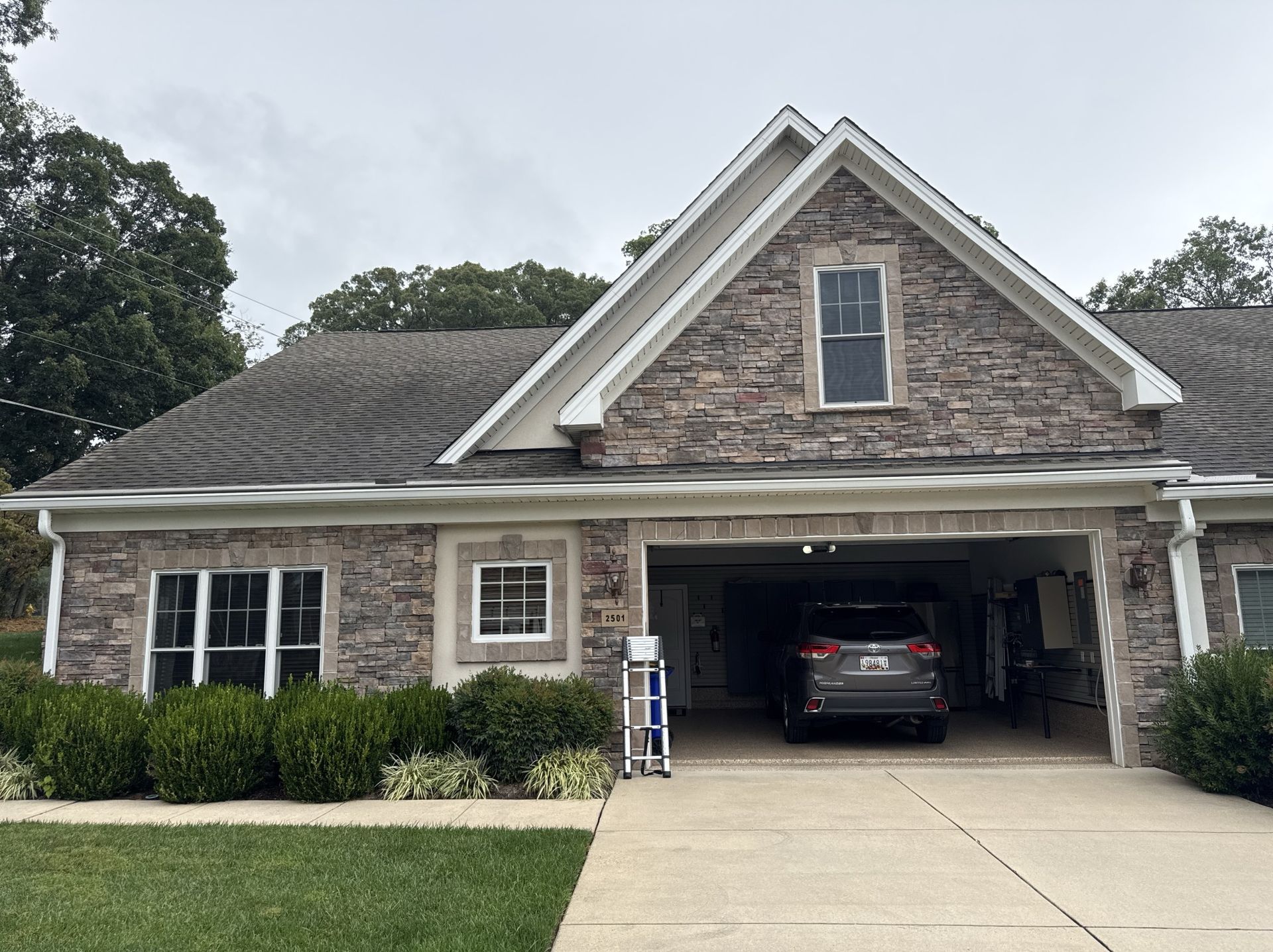 Stone-faced house with a garage, gray car inside, and shrubs in front on a cloudy day.