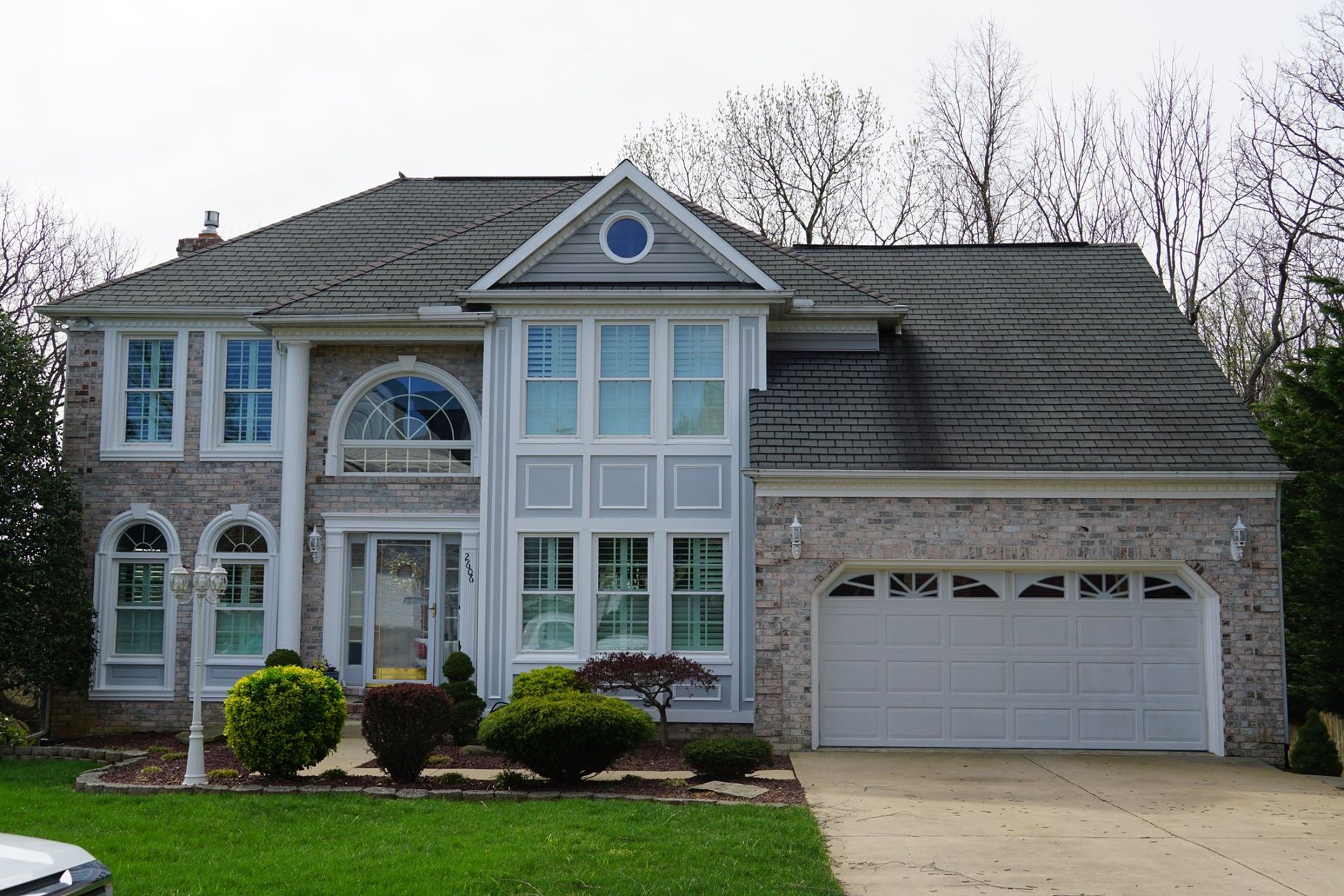 Two-story brick house with white trim, arched windows, and a two-car garage. Lush green lawn and cloudy sky.