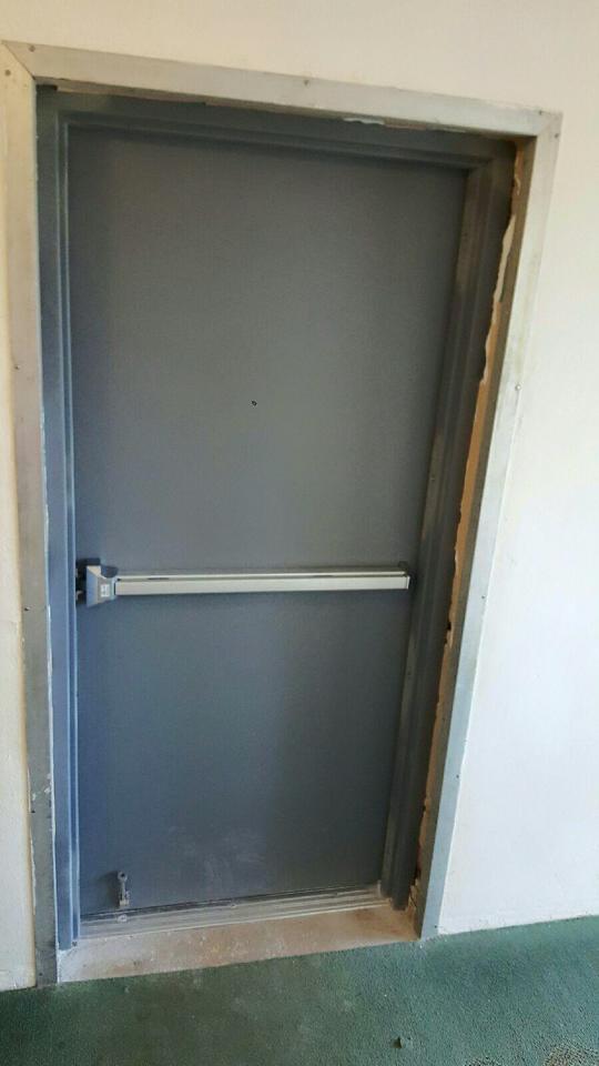 A gray door with a panic bar on it is in a hallway.