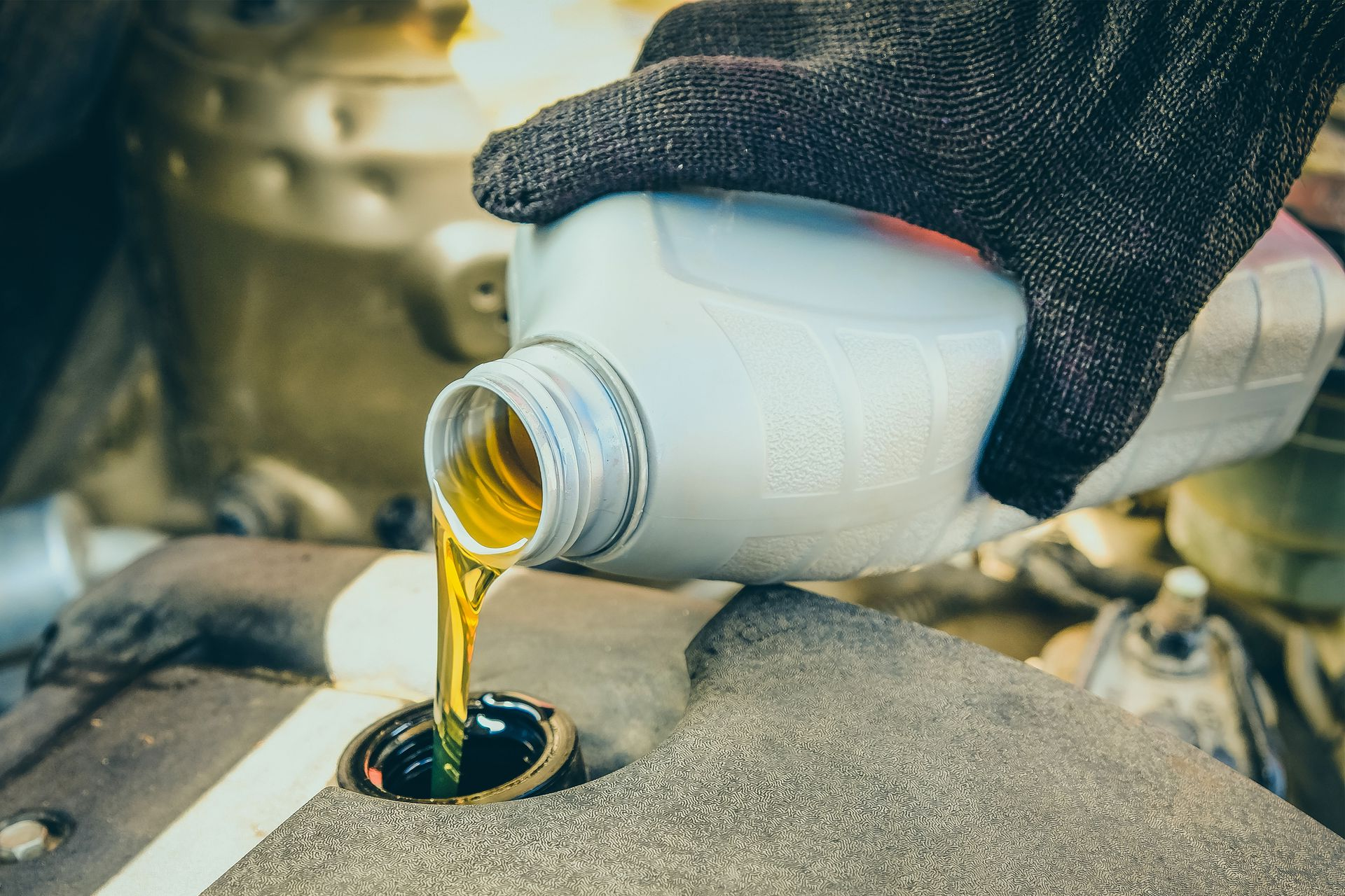Gloved hand pouring golden oil from a white container into an engine fill opening