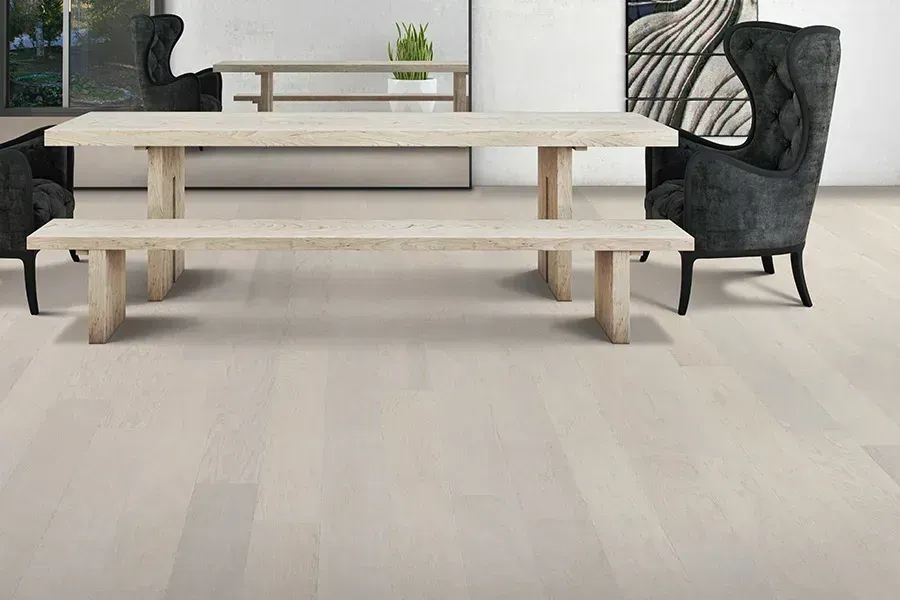 Bright dining room with a long wooden table, bench seating, and a dark upholstered chair on light flooring.