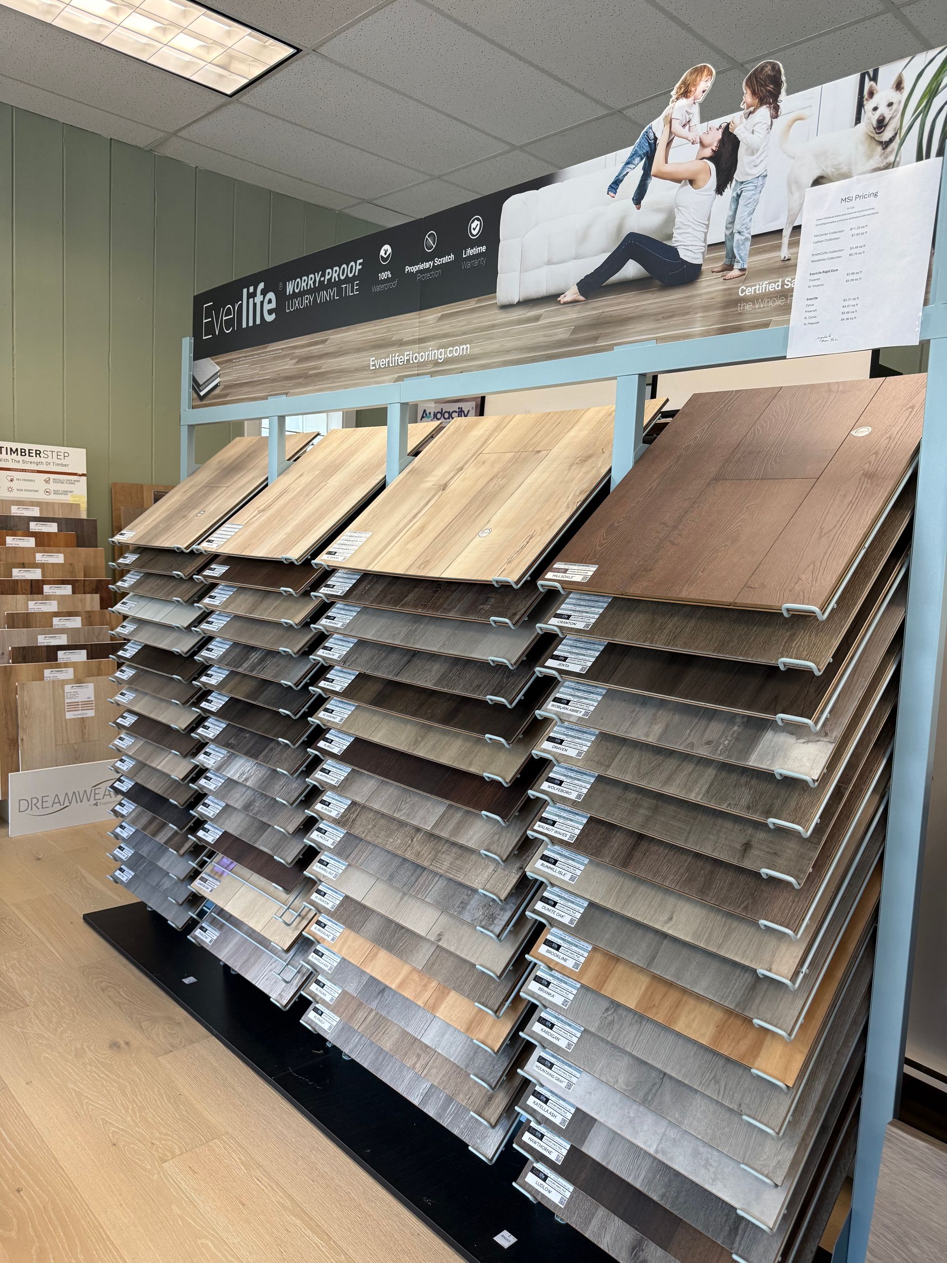 Tile display rack in a store aisle with rows of flooring samples in neutral tones and a wall sign above.