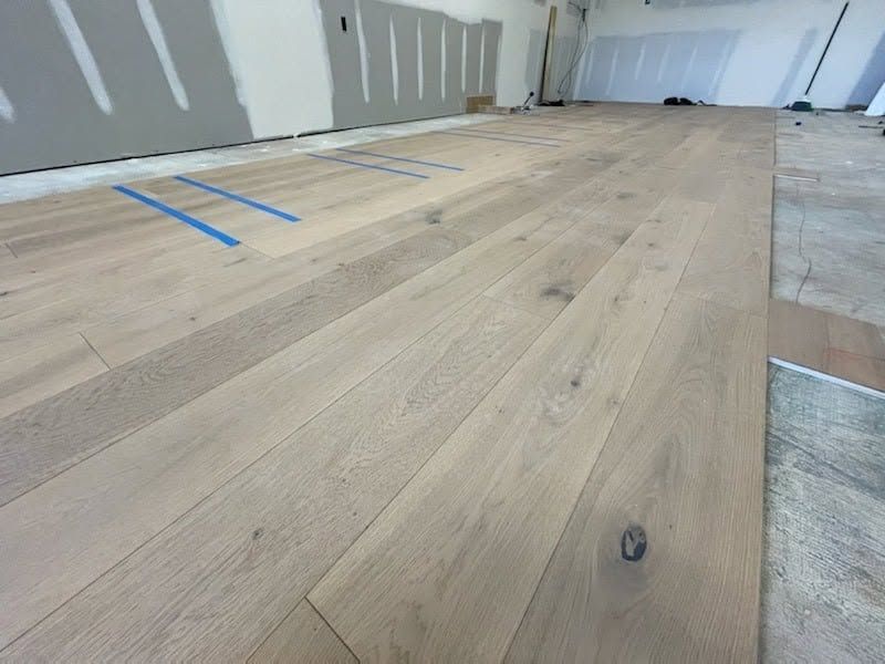 Light wood flooring under construction in an unfinished room with drywall and blue tape markings.