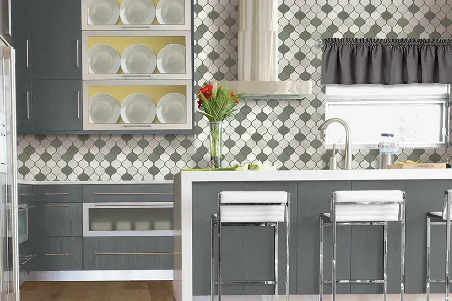 Modern kitchen with gray cabinets, patterned backsplash, island stools, and decorative dish shelves