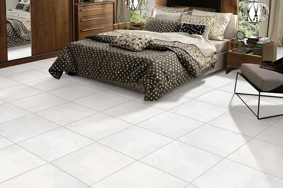 Modern bedroom with a patterned bedspread, white tiled floor, wooden dresser, and lounge chair