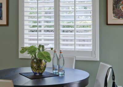 A round dark table with a glass vase of greenery and two water bottles in front of a window with white plantation shutters.