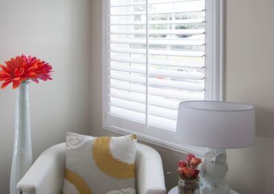 A room corner with a white shuttered window, a white chair with a yellow patterned pillow, a tall vase of red flowers, and lamp.