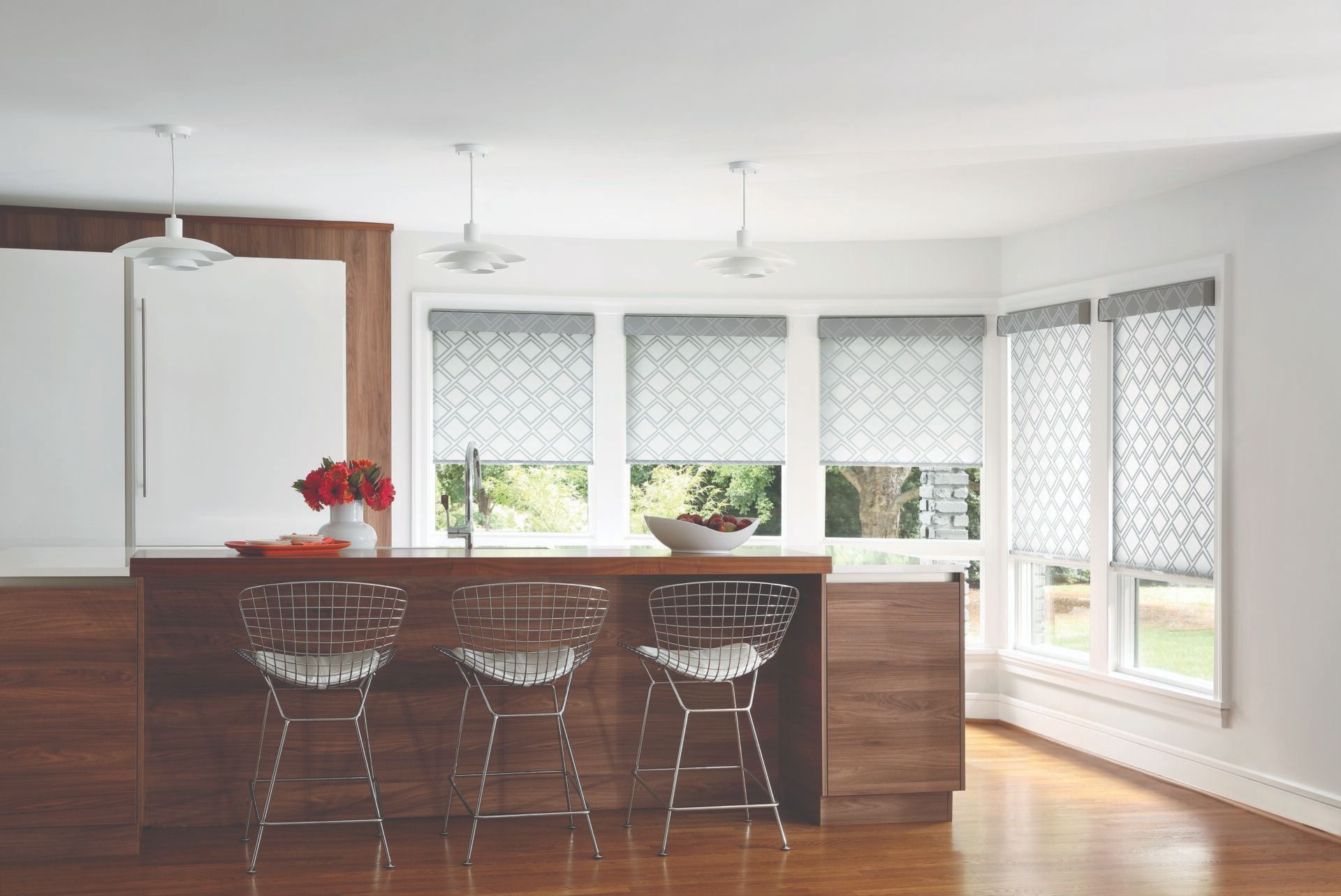 Modern kitchen with a wood-paneled island, three wire stools, and windows covered with patterned roller shades.