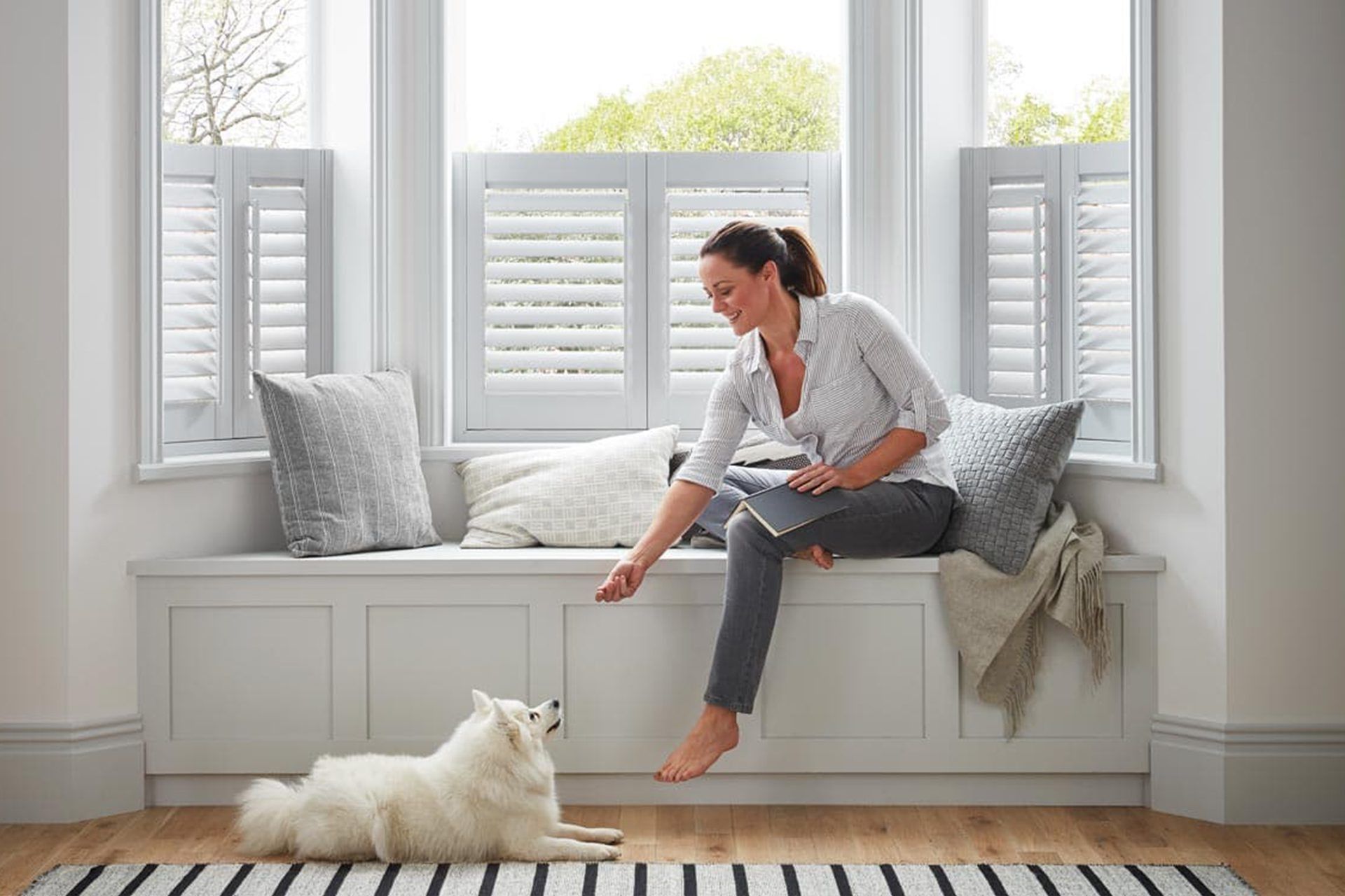 A person sits on a window seat and interacts with a small white dog in a brightly lit room with white shutters.