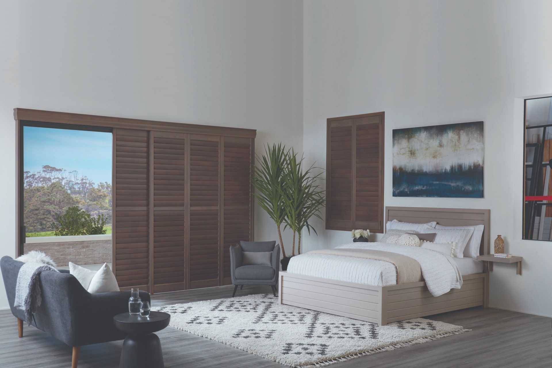 A modern, airy bedroom features a bed, armchair, and couch, decorated with dark wood shutters and a neutral area rug.