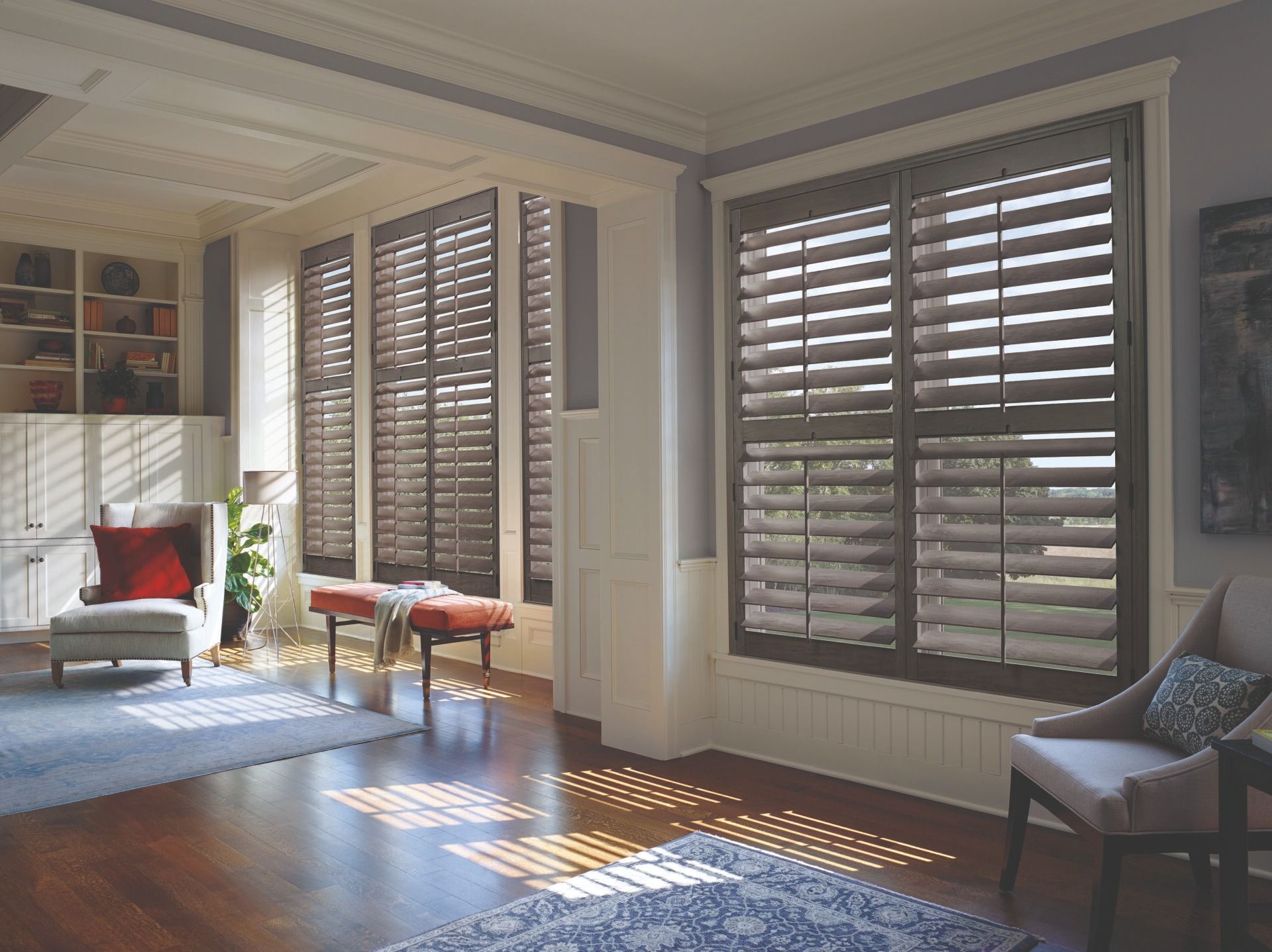 Sunlight streams through dark brown plantation shutters into a room with hardwood floors, chairs, and a bench.