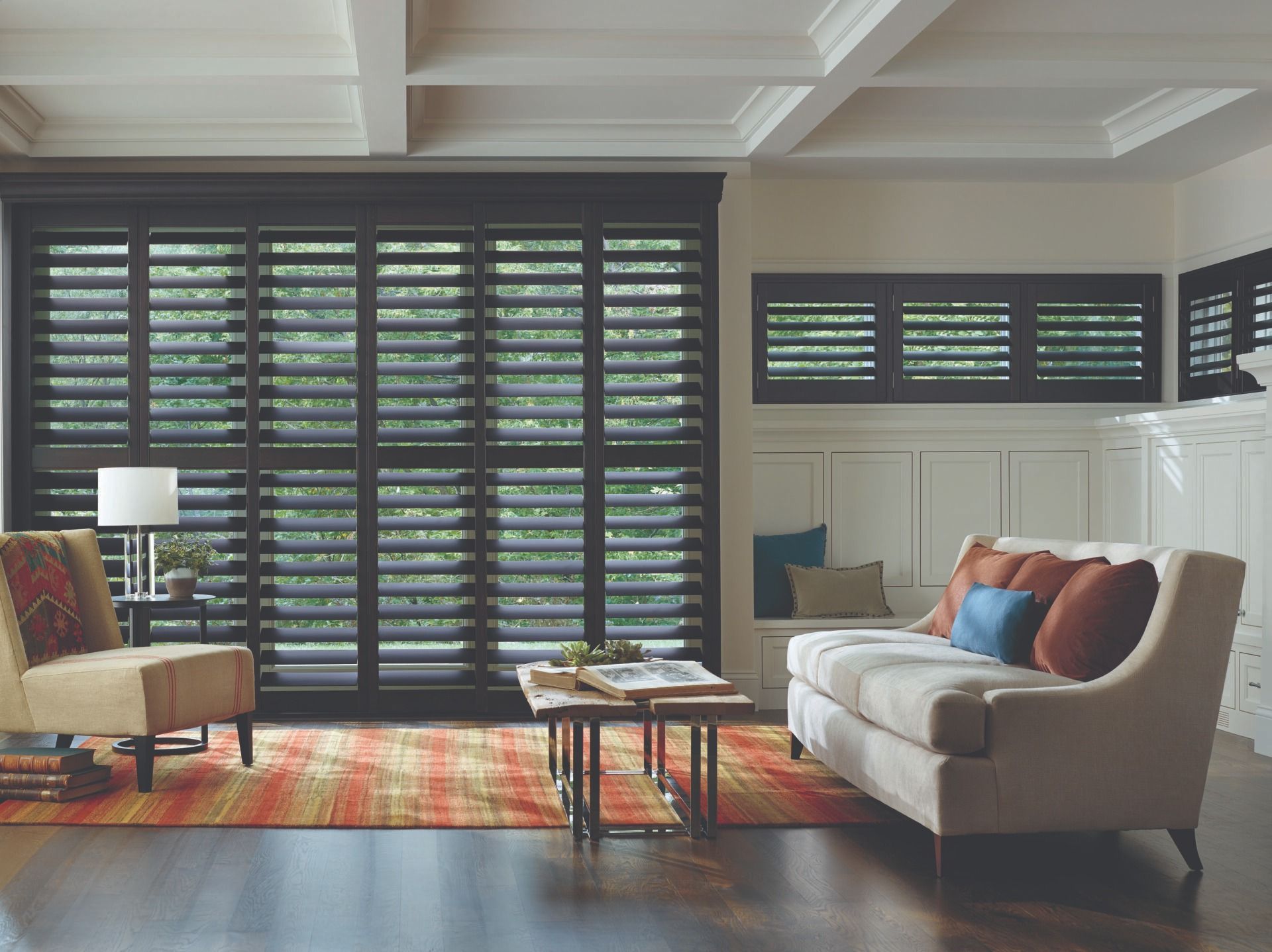 A brightly lit living room with dark wooden shutter walls, a sofa, a patterned rug, and a chair by a lamp.