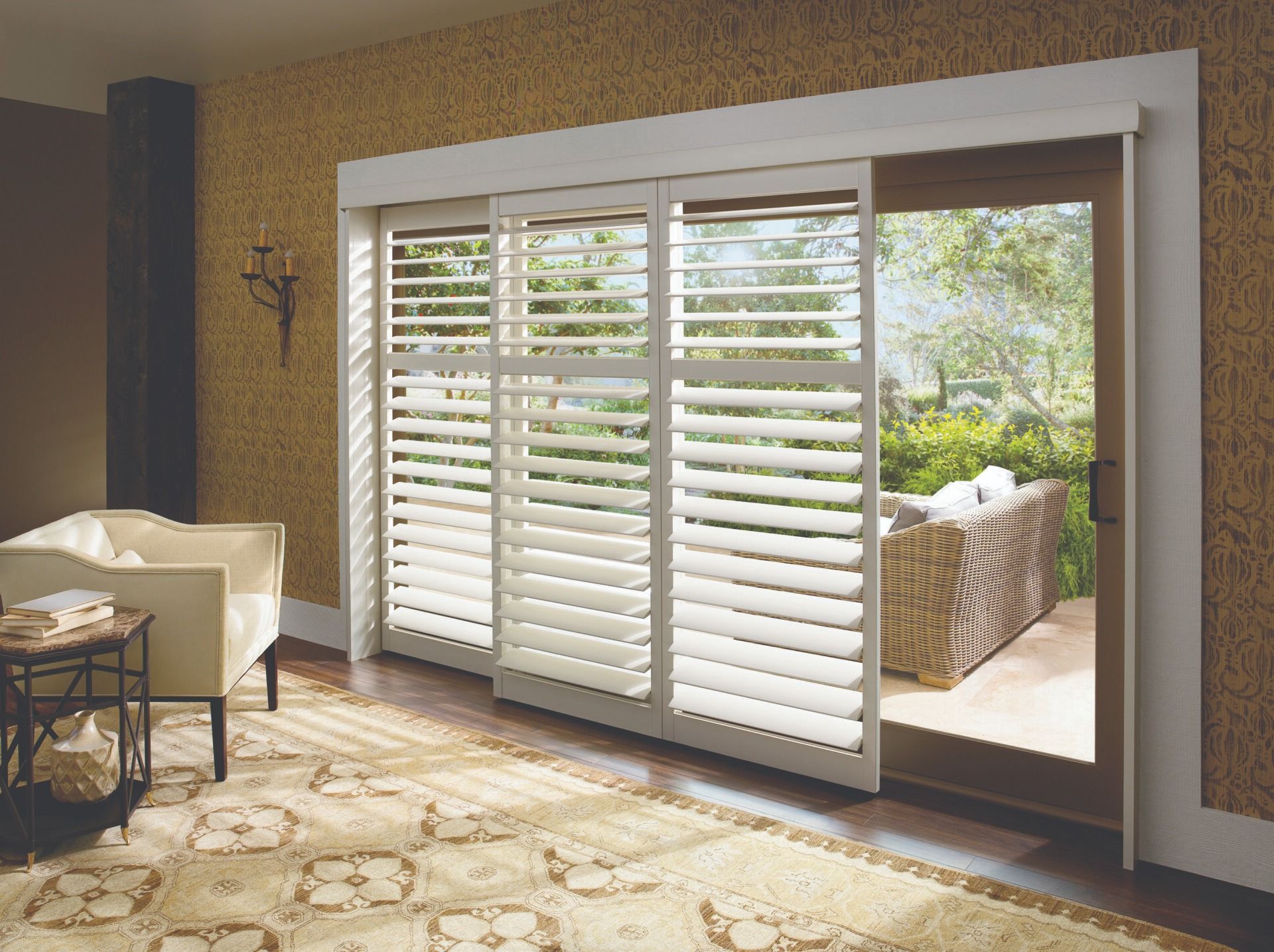 White plantation shutters installed on a sliding glass door in a cozy, sunlit living room with patterned rug and armchair.