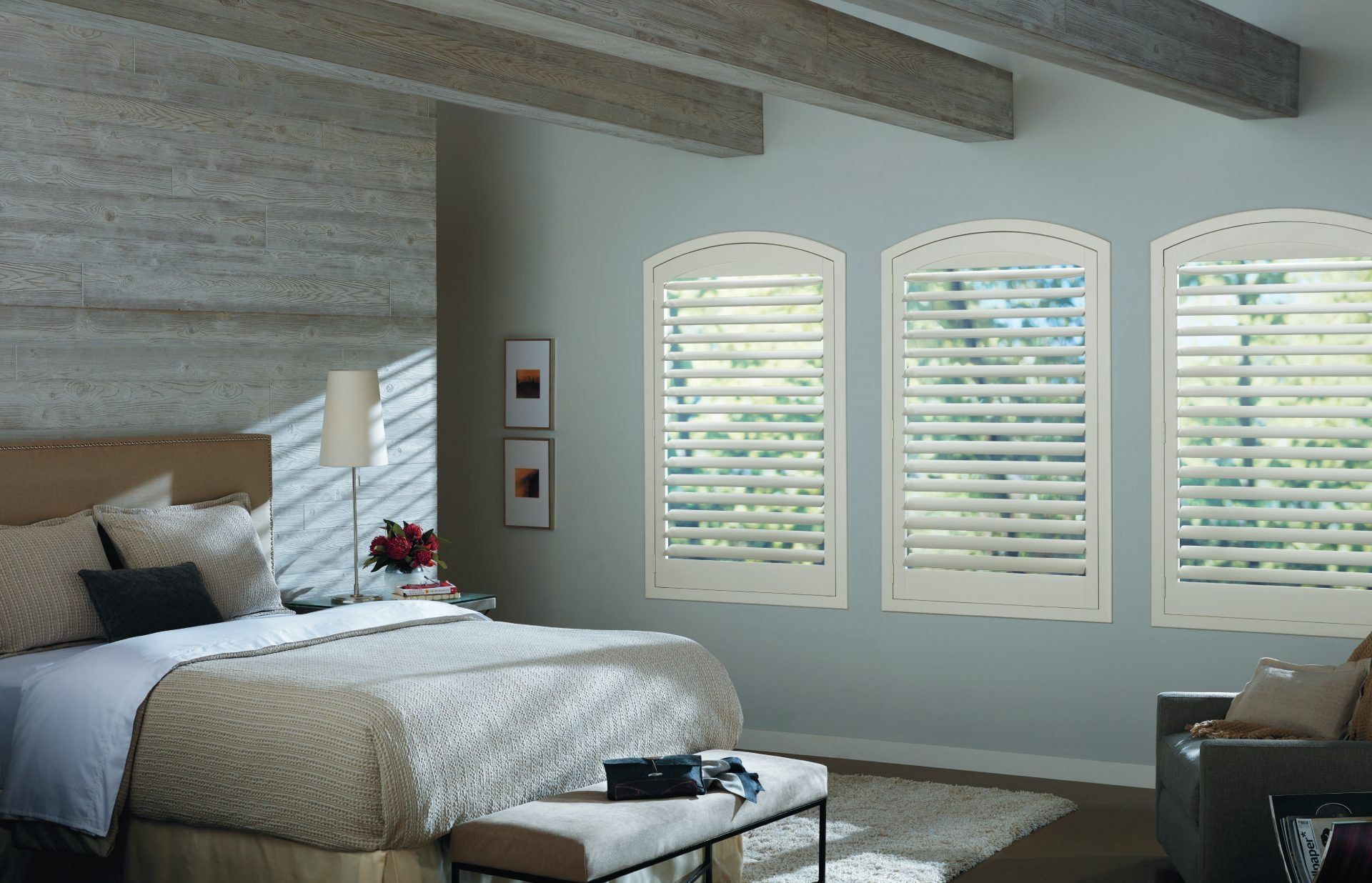 A bedroom with a queen bed, wooden ceiling beams, and three arched windows covered with white plantation shutters.