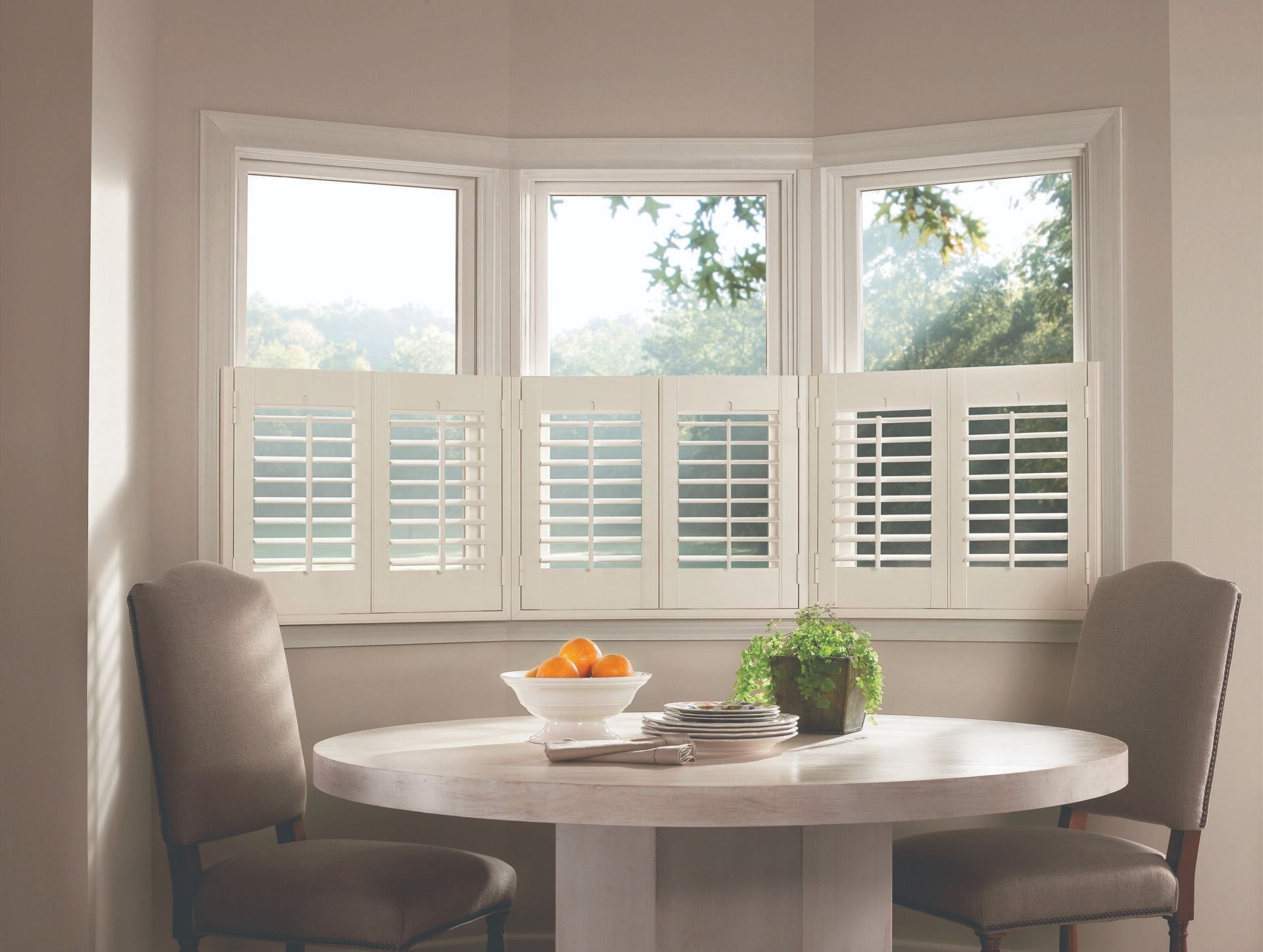 A dining nook with a round table, two chairs, and a bay window featuring white plantation shutters.