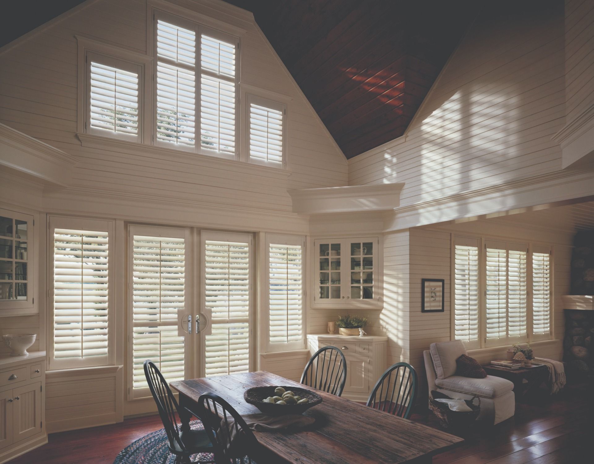 A high-ceilinged room with a wooden table, chairs, and multiple windows covered by white plantation shutters.