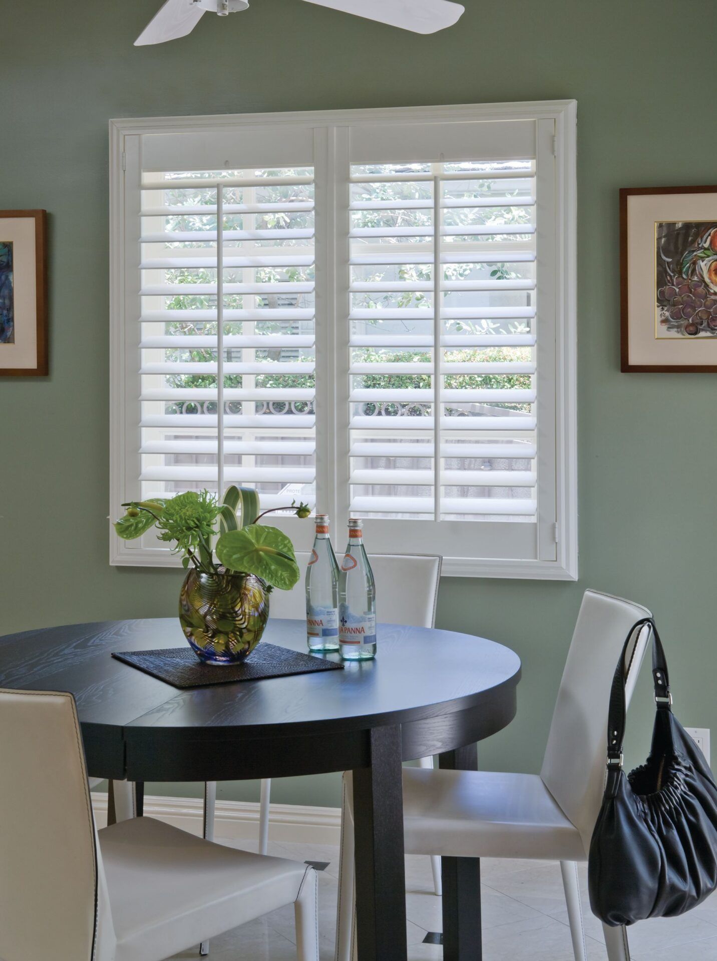 A round black dining table with chairs and a plant sits in front of a window covered by white plantation shutters.