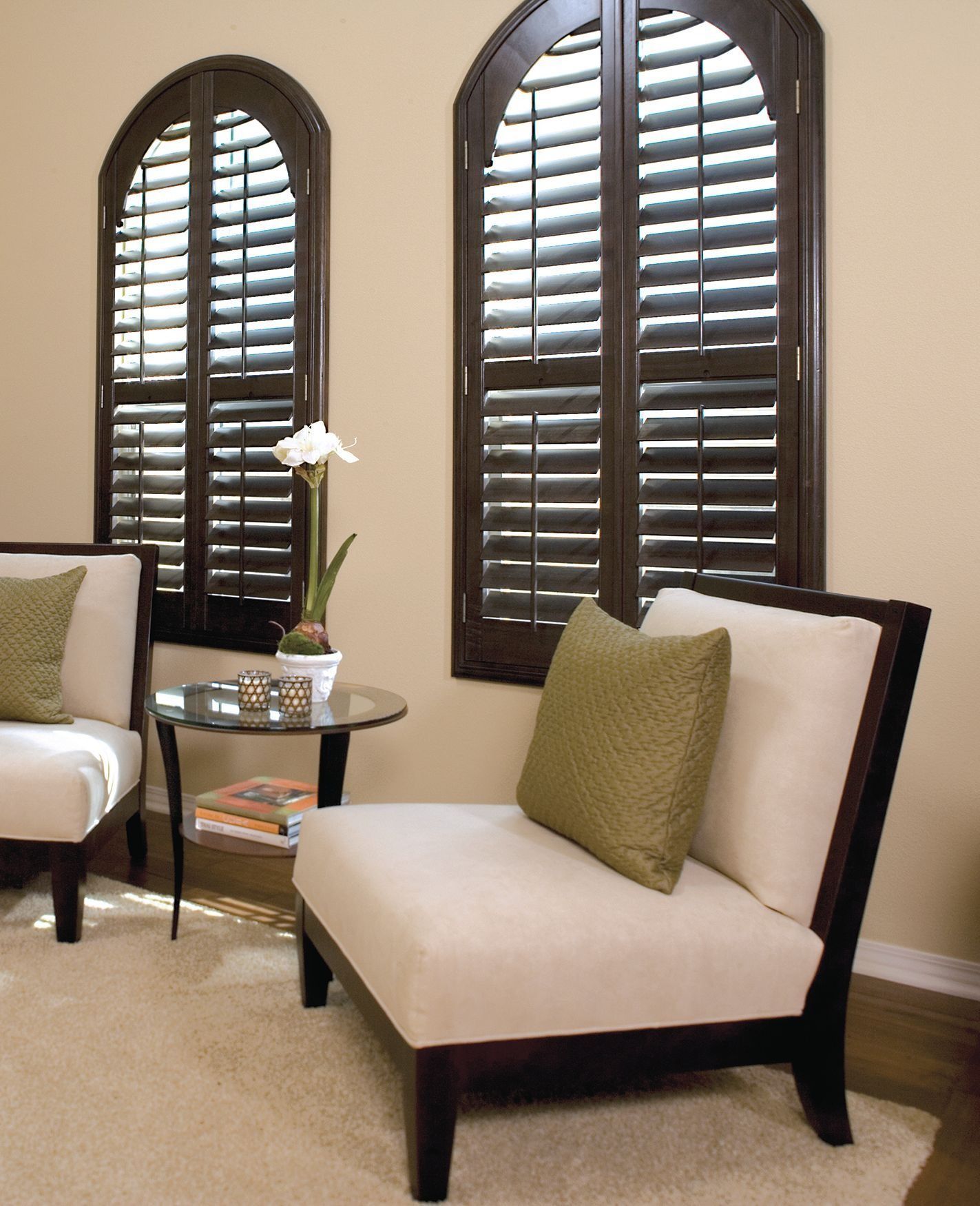 Two dark wooden arched shutters cover windows in a room with a side table, a plant, and two off-white chairs.