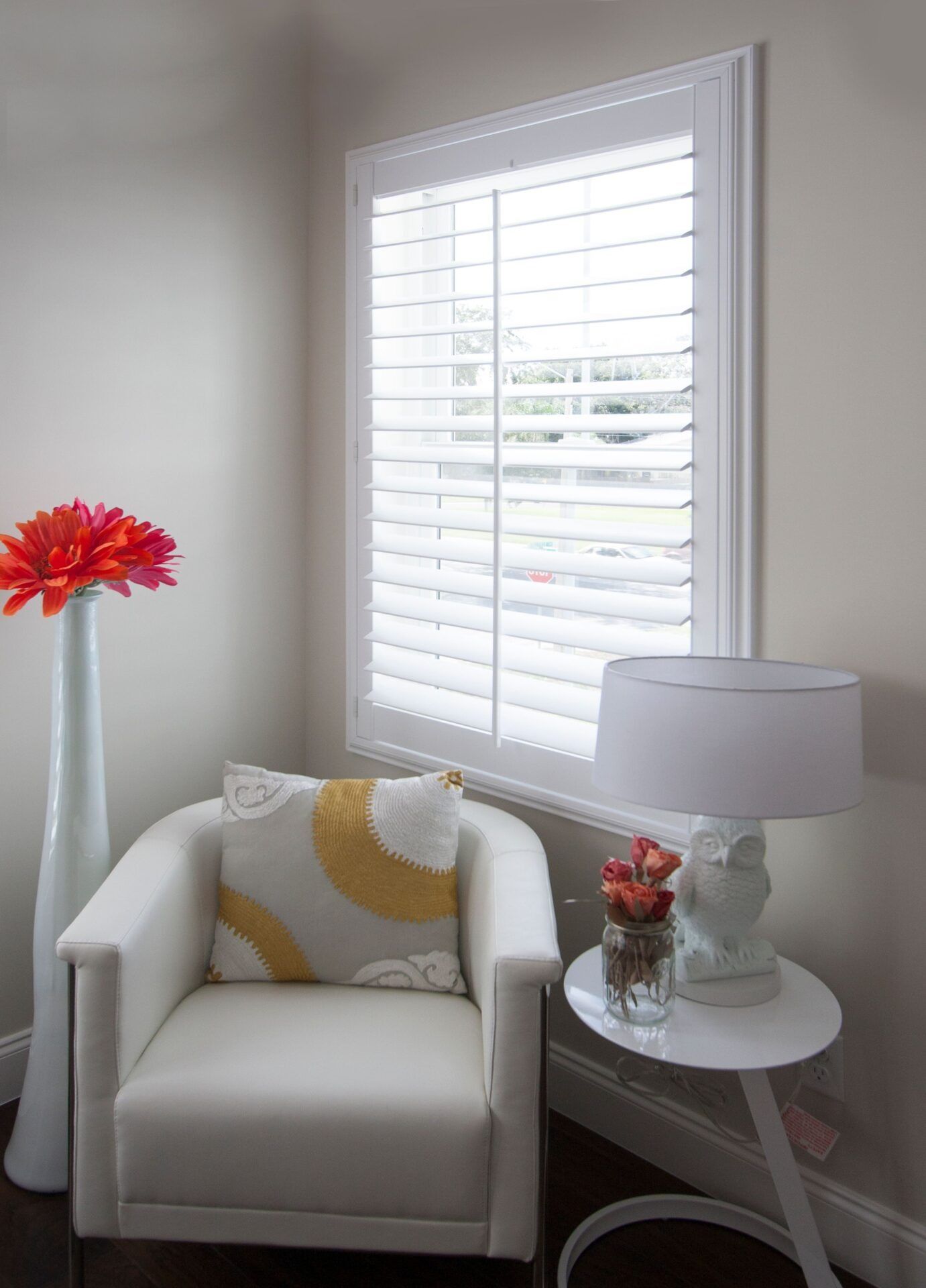 A white armchair with a patterned pillow, a white lamp on a side table, and a tall floor vase with flowers by a window.