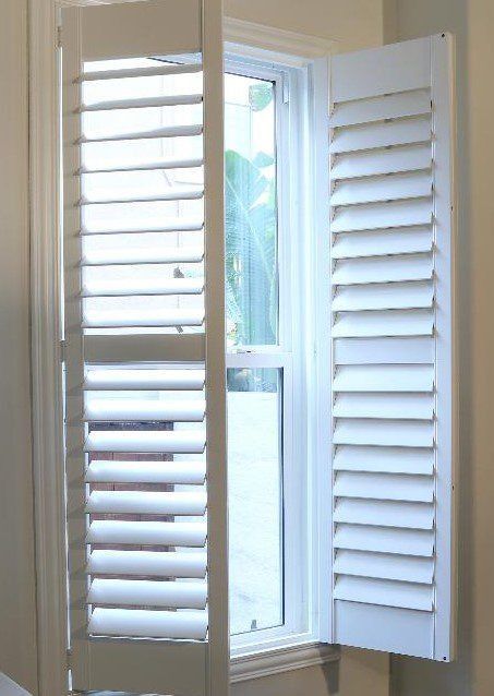 White plantation-style window shutters are open, revealing a view of outdoor greenery through the windowpane.