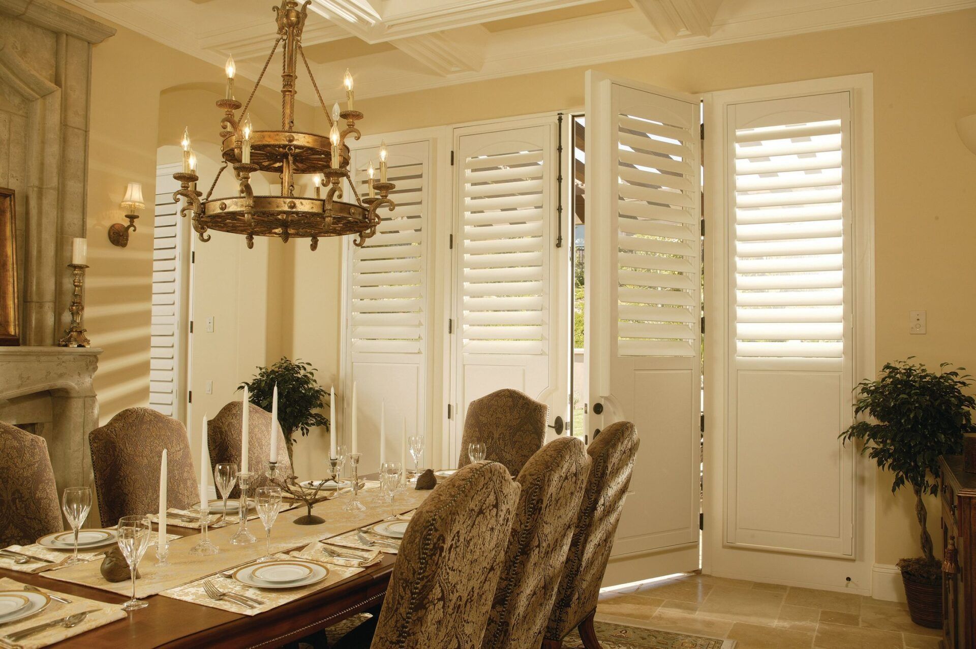 A dining room with a long table, chandelier, and white plantation shutter doors opening to an outdoor space.