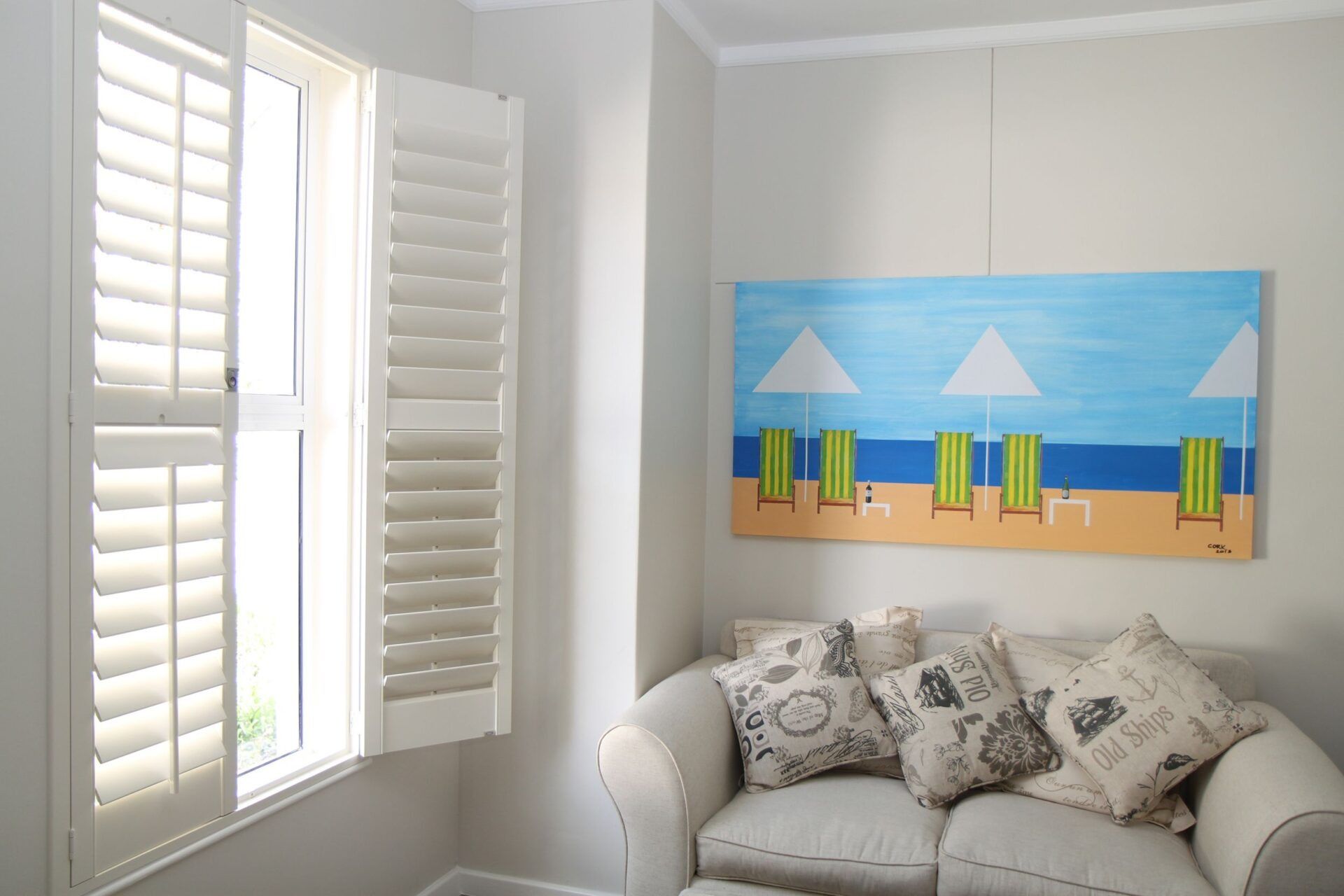 A window with white shutters next to a small beige sofa featuring patterned pillows and a beach-themed wall painting.