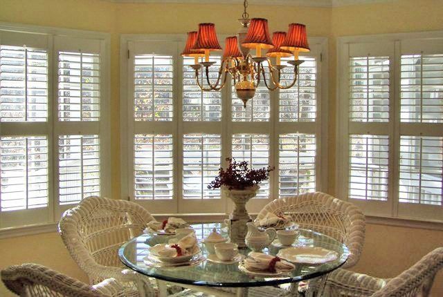 A glass-top dining table with wicker chairs sits in a sunlit bay window under a bronze chandelier with red shades.