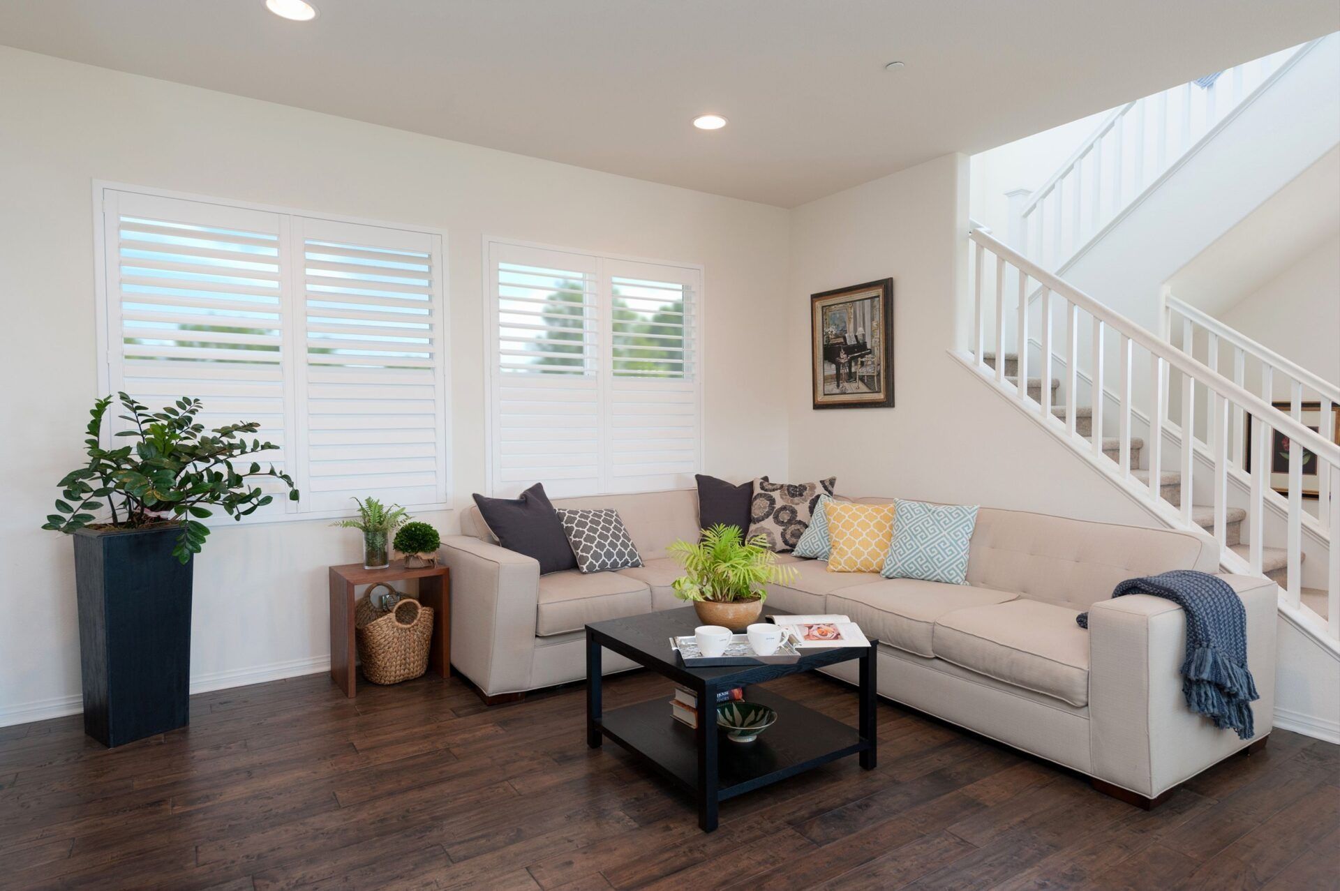 A modern living room with a beige sectional sofa, a black coffee table, a houseplant, and a staircase.
