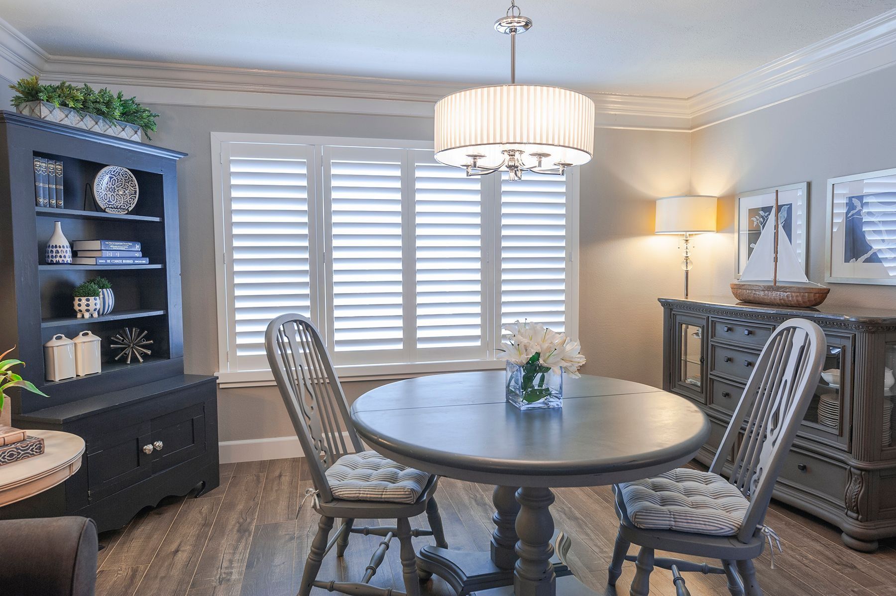 A dining room with a round gray table, two gray chairs, a dark hutch, and large white plantation shutters on the windows.