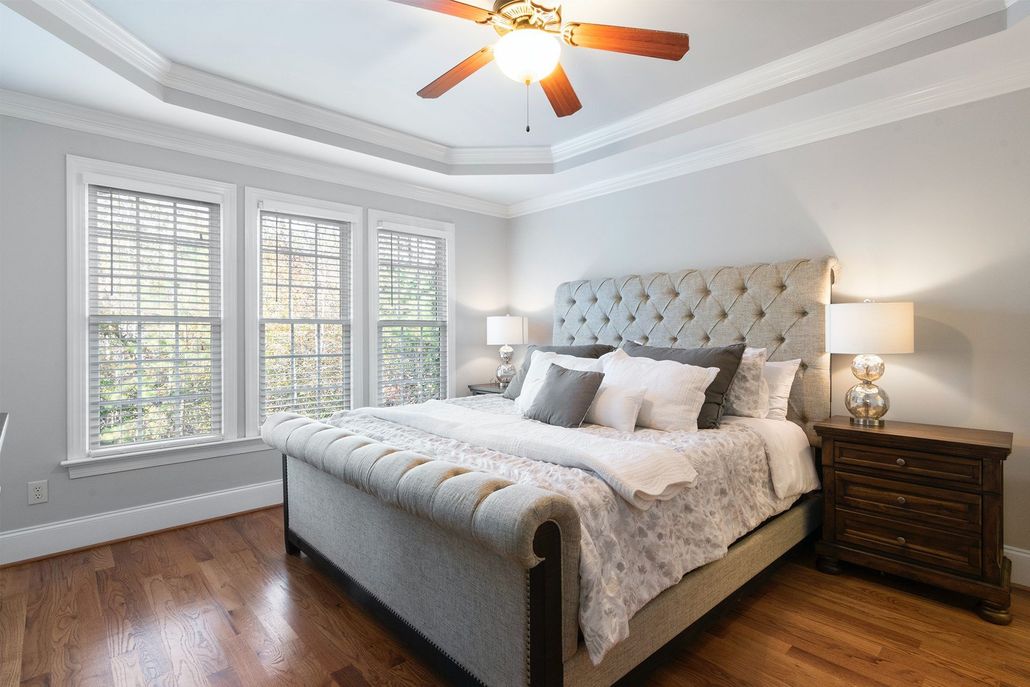A modern bedroom with a tufted gray bed, white bedding, large windows, hardwood floors, and a ceiling fan.