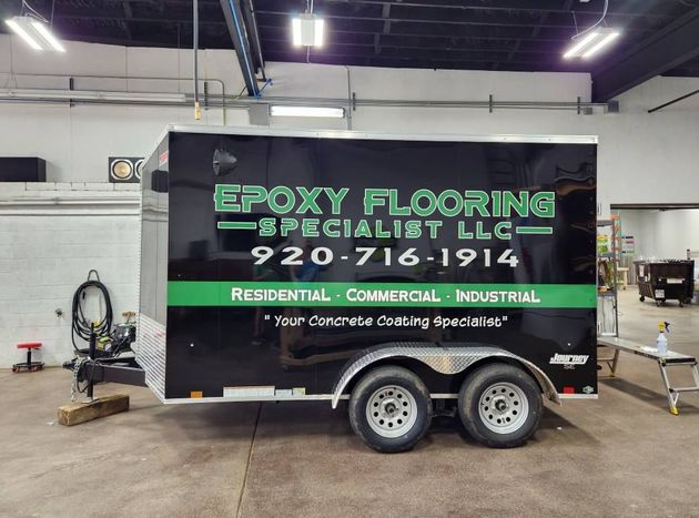 A black cargo trailer for Epoxy Flooring Specialist LLC parked indoors, featuring green lettering and a phone number.