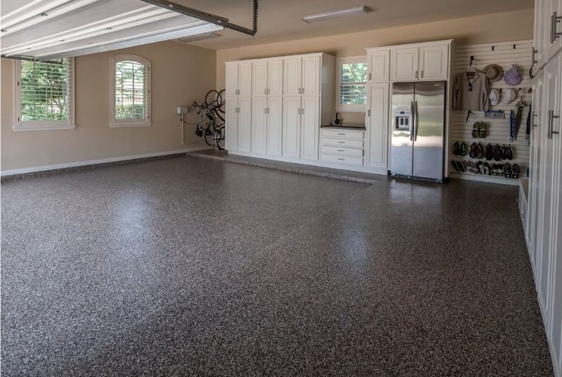 A spacious, tidy garage with grey speckled floors, white cabinetry, a stainless steel refrigerator, and wall storage.