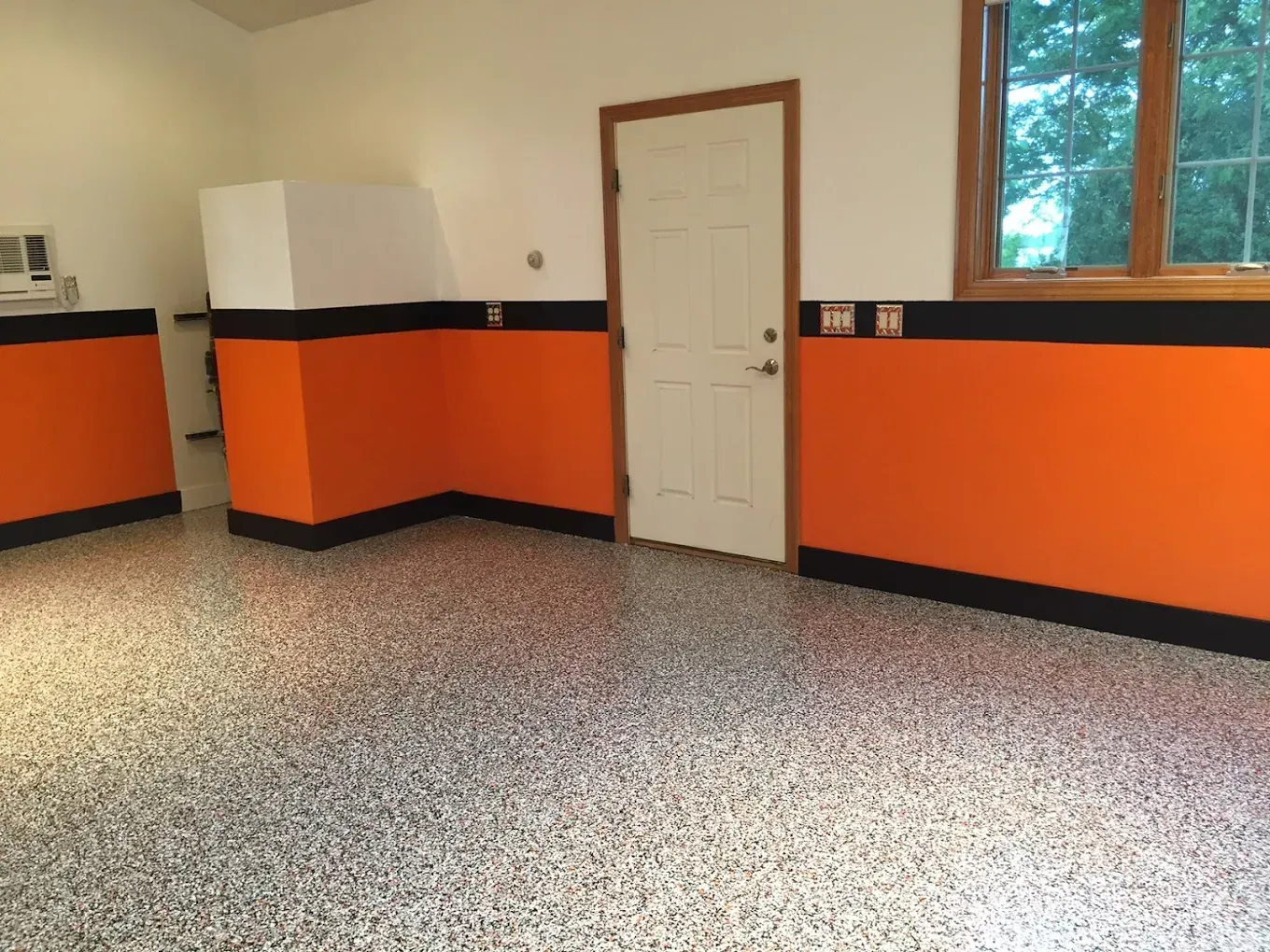 A garage interior with gray speckled flooring, orange walls with black trim, a white door, and a wooden window frame.