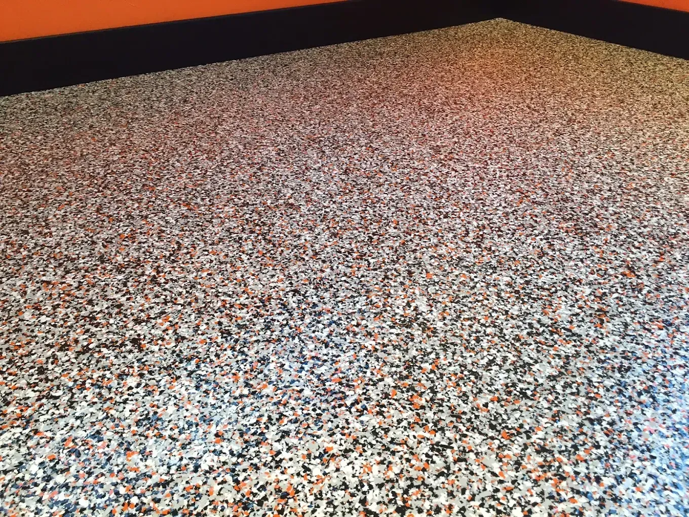 A close-up of a speckled, multi-colored epoxy floor with a black baseboard against an orange wall.